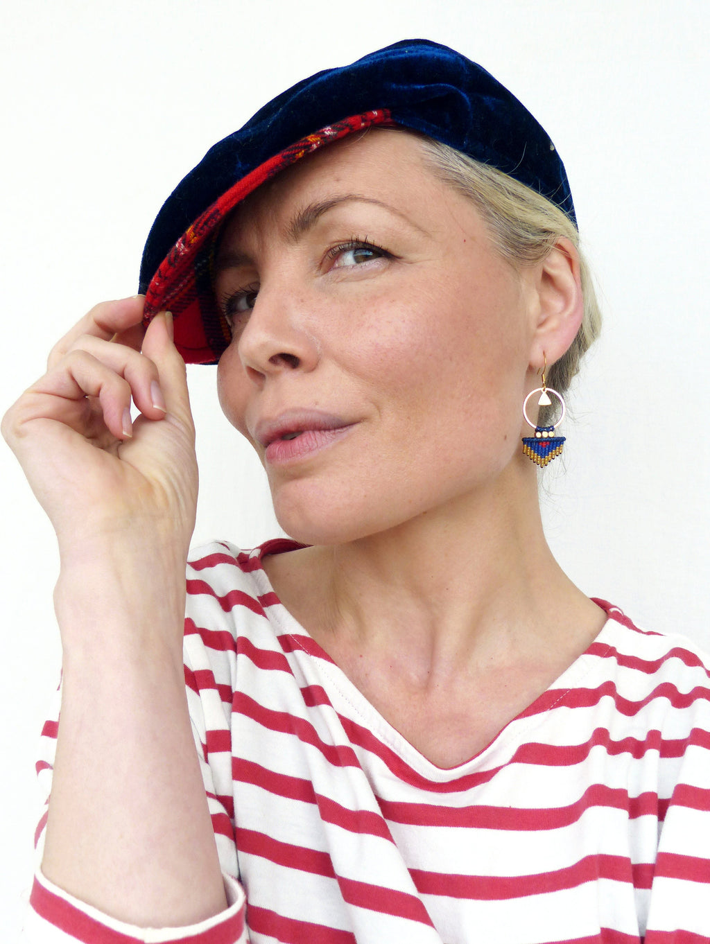 Peut inclure: Une femme portant une chemise à rayures rouges et bleues et une casquette en velours bleu avec une doublure à motifs rouges et blancs. Elle porte des boucles d'oreilles en perles dorées et bleues.
