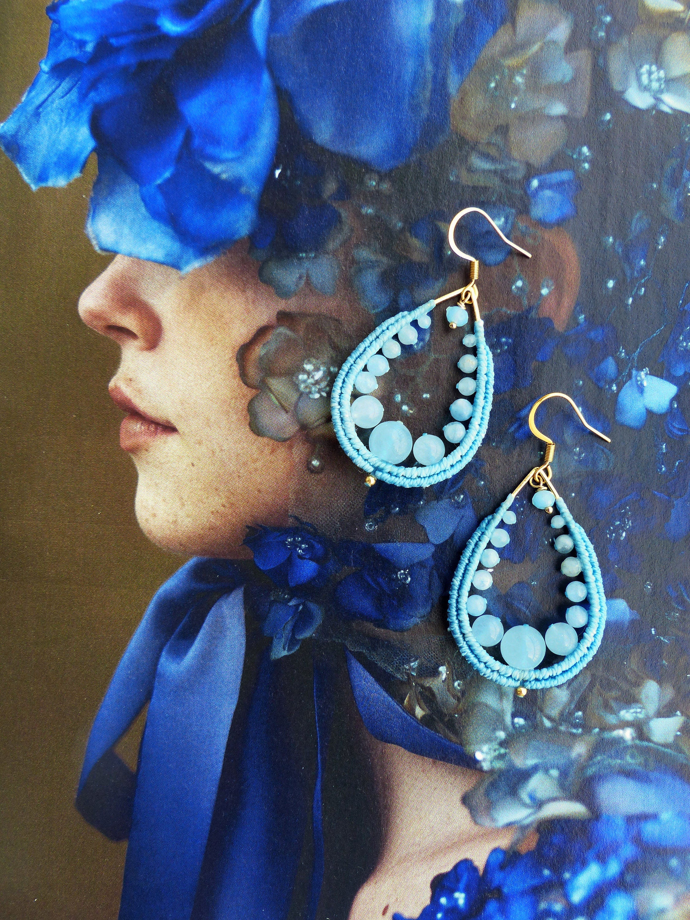 Peut inclure: Une paire de boucles d'oreilles en forme de larme avec des perles bleues et des accents dorés. Les boucles d'oreilles pendent de l'oreille d'un mannequin.
