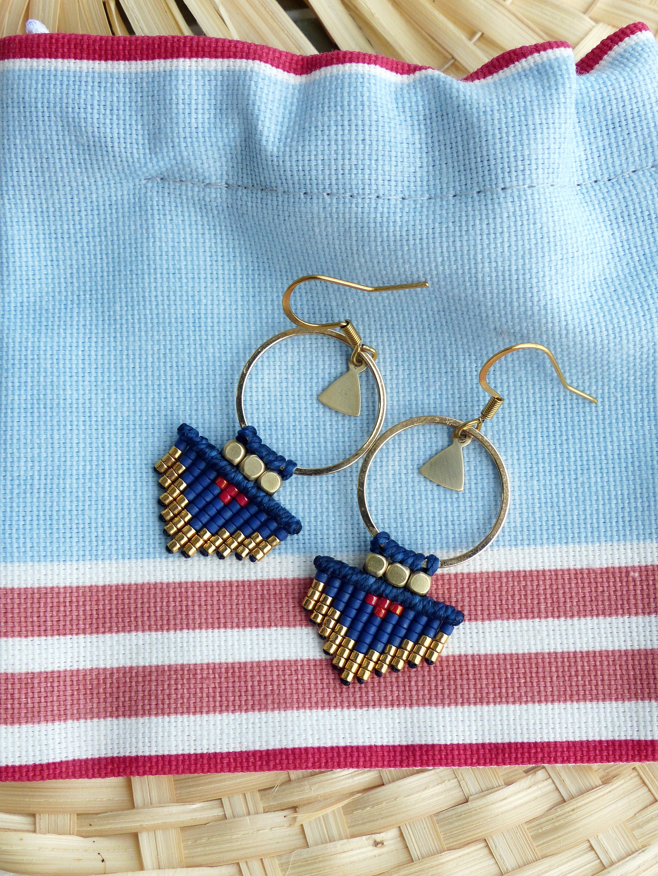 Peut inclure: Une paire de boucles d'oreilles en perles bleues et dorées avec un charme triangulaire suspendu à un anneau doré. Les boucles d'oreilles sont sur un fond de tissu rayé bleu et blanc.