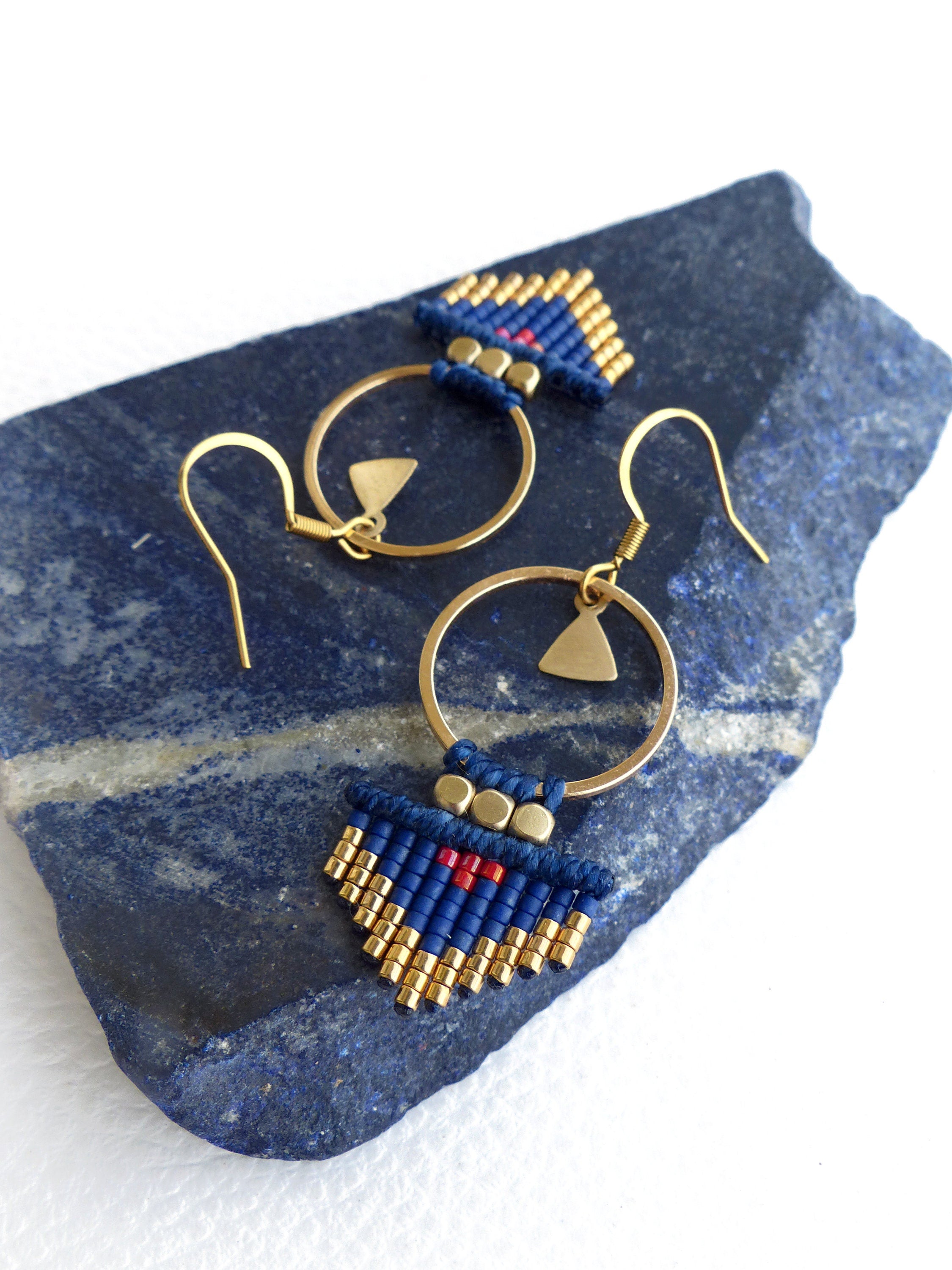 Peut inclure: Une paire de boucles d'oreilles créoles dorées avec des franges de perles bleues et dorées et un accent rouge. Les boucles d'oreilles sont suspendues à une pierre bleue.