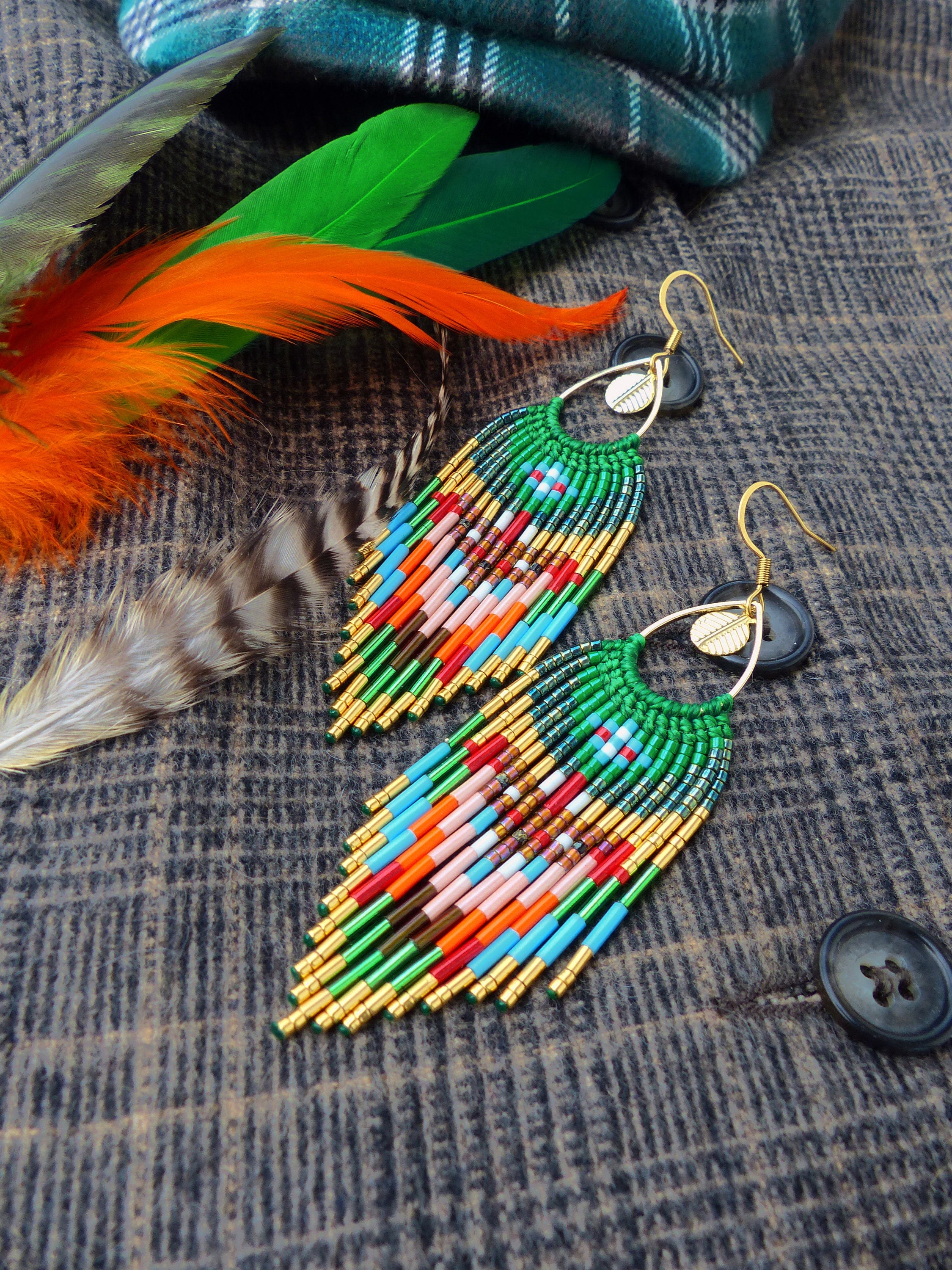 Peut inclure: Une paire de longues boucles d'oreilles en perles avec un schéma de couleurs vert, bleu, orange et or. Les boucles d'oreilles sont suspendues à un fond en tissu texturé.