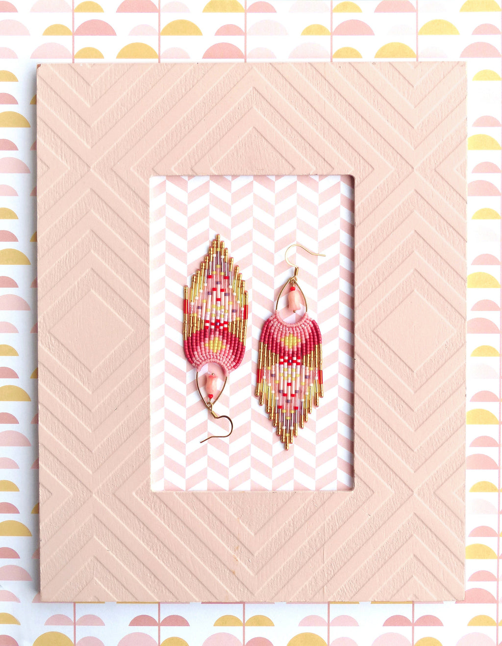 Peut inclure: Une paire de boucles d'oreilles en perles roses et dorées avec un design en frange. Les boucles d'oreilles sont suspendues à un fond rose avec un motif géométrique.