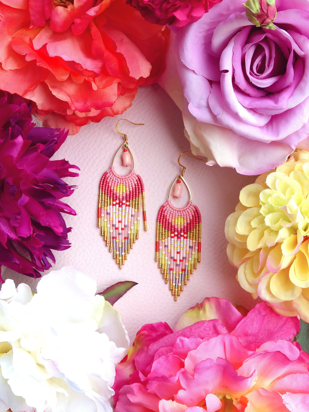 Peut inclure: Une paire de boucles d'oreilles en perles roses, jaunes et dorées avec un accent de pierre rose. Les boucles d'oreilles sont suspendues à des crochets dorés et sont entourées de fleurs roses, violettes et blanches.