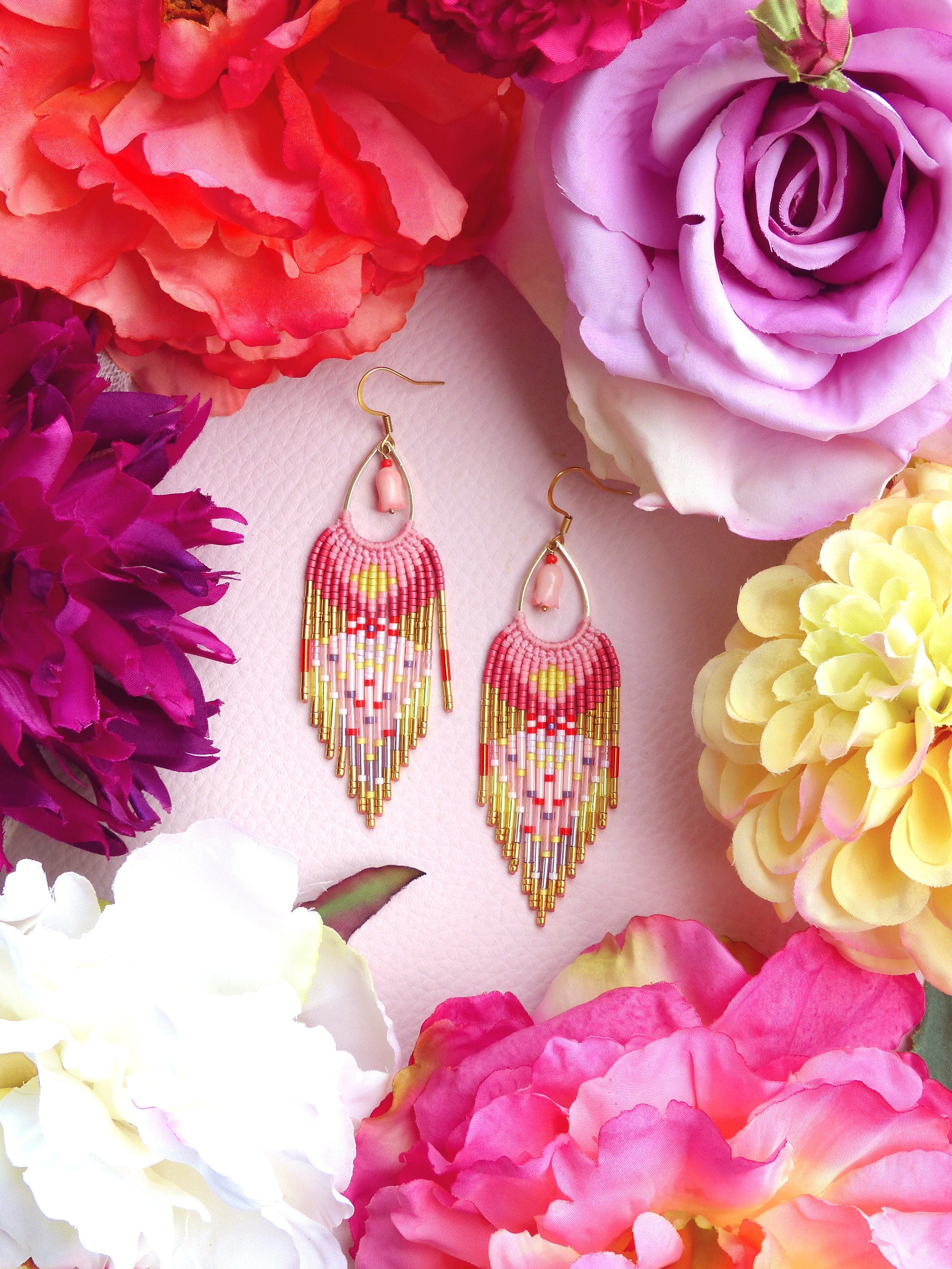 Peut inclure: Une paire de boucles d'oreilles en perles roses, jaunes et dorées avec un accent de pierre rose. Les boucles d'oreilles sont suspendues à des crochets dorés et sont entourées de fleurs roses, violettes et blanches.