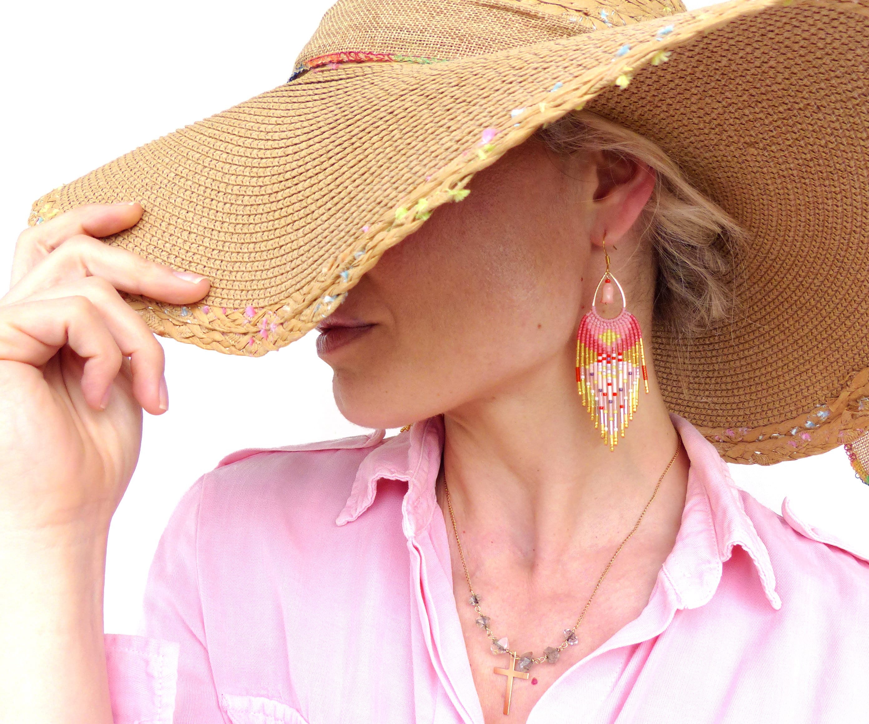 Peut inclure: Une femme portant un chapeau de paille avec des accents colorés et une paire de boucles d'oreilles en perles roses et dorées. Elle porte une chemise rose et un collier en or avec un pendentif en forme de croix.