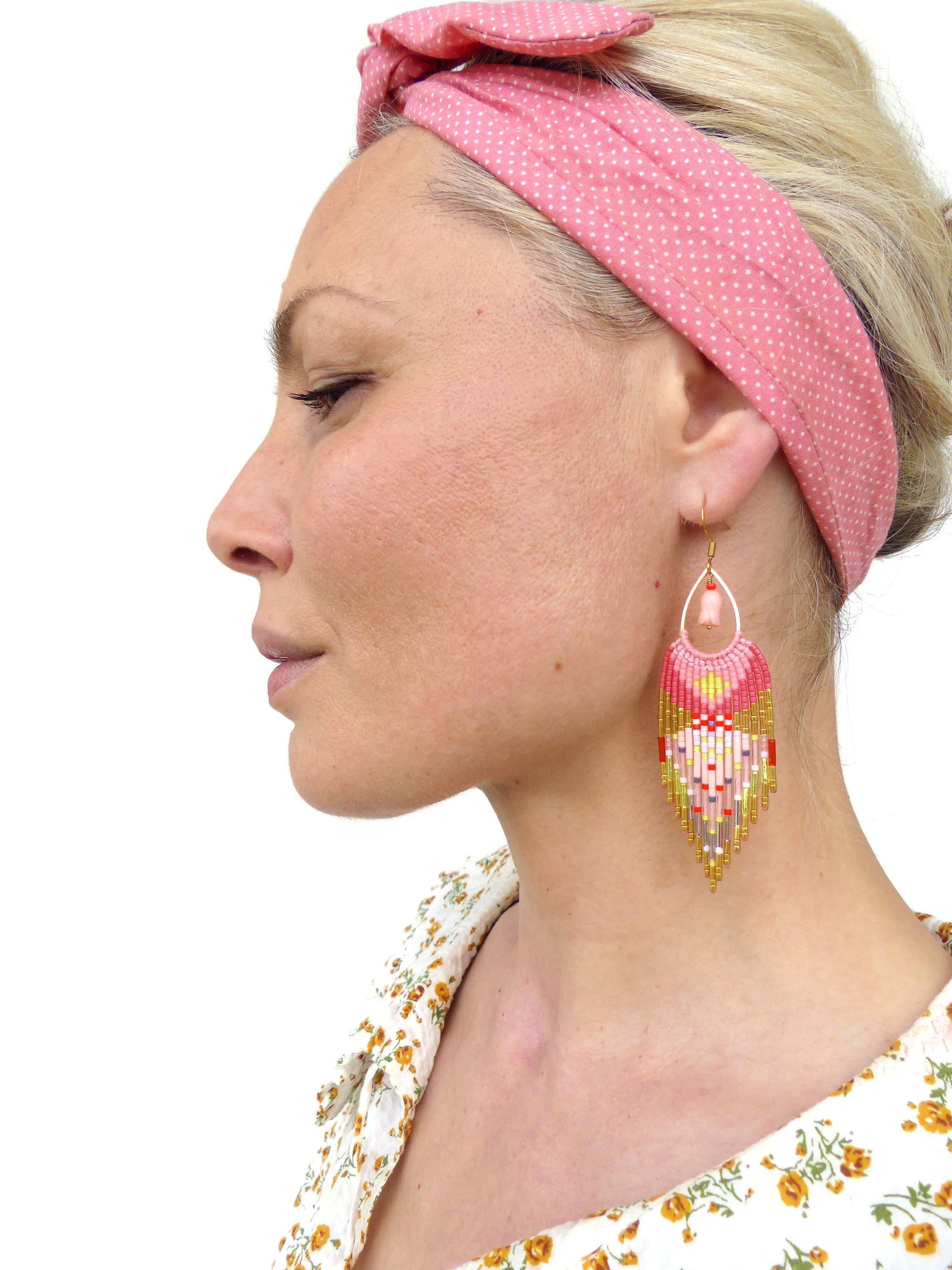 Peut inclure: Une paire de boucles d'oreilles en perles roses, dorées et blanches avec un design en frange. Les boucles d'oreilles pendent des oreilles d'une personne portant un bandeau rose.