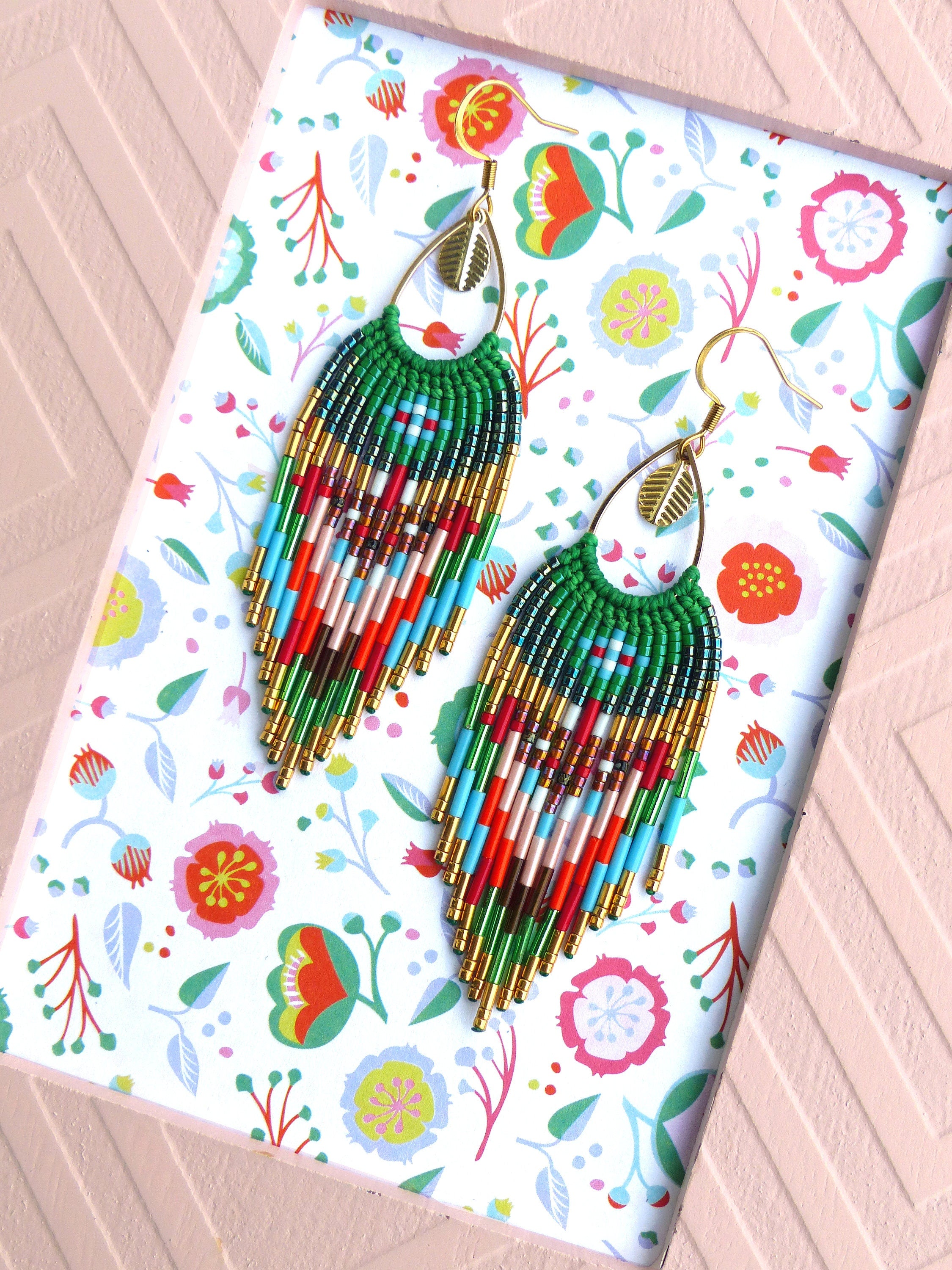 Peut inclure: Une paire de longues boucles d'oreilles en perles avec un motif de couleurs vert, bleu, rouge et or. Les boucles d'oreilles sont suspendues à des crochets dorés et ont un charme en forme de feuille.