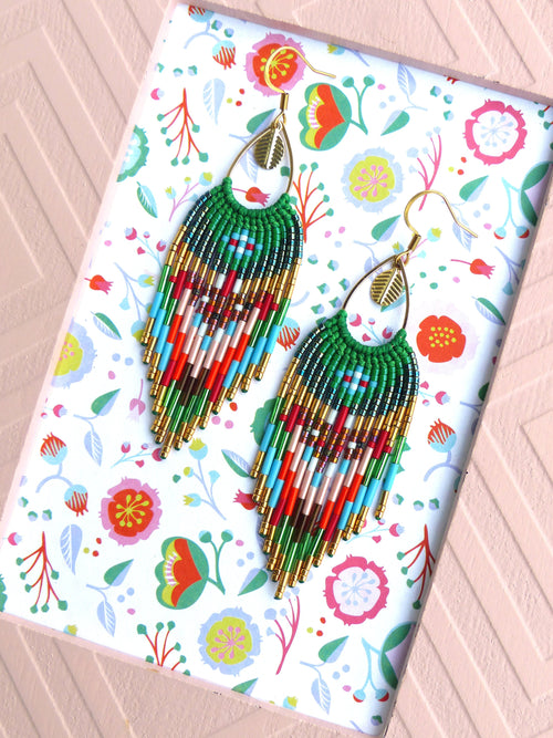 Peut inclure: Une paire de longues boucles d'oreilles en perles avec un motif de couleurs vert, bleu, rouge et or. Les boucles d'oreilles sont suspendues à des crochets dorés et ont un charme en forme de feuille.