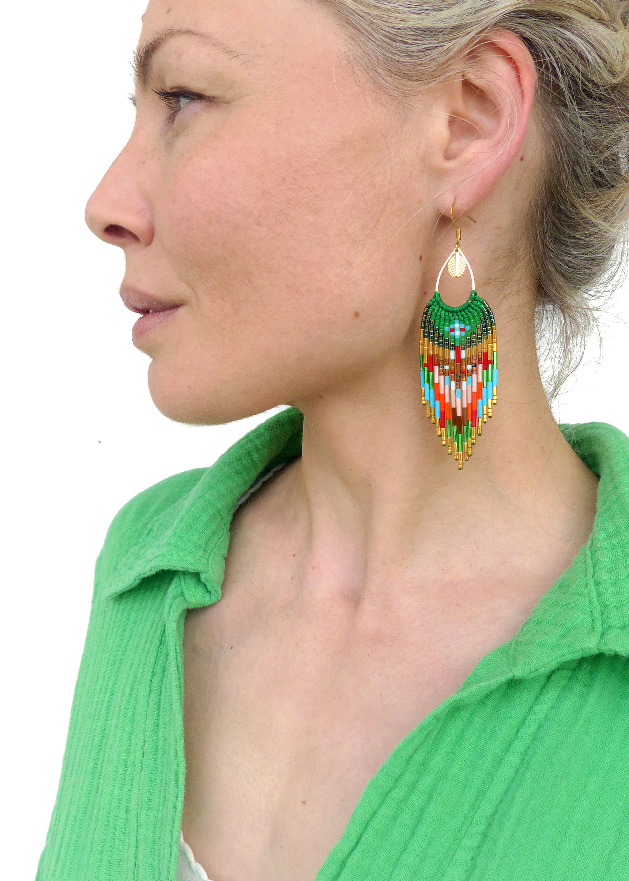 Peut inclure: Une paire de longues boucles d'oreilles vertes en perles avec des accents dorés et des motifs géométriques colorés. Les boucles d'oreilles pendent des lobes d'oreilles d'une personne portant une chemise verte.