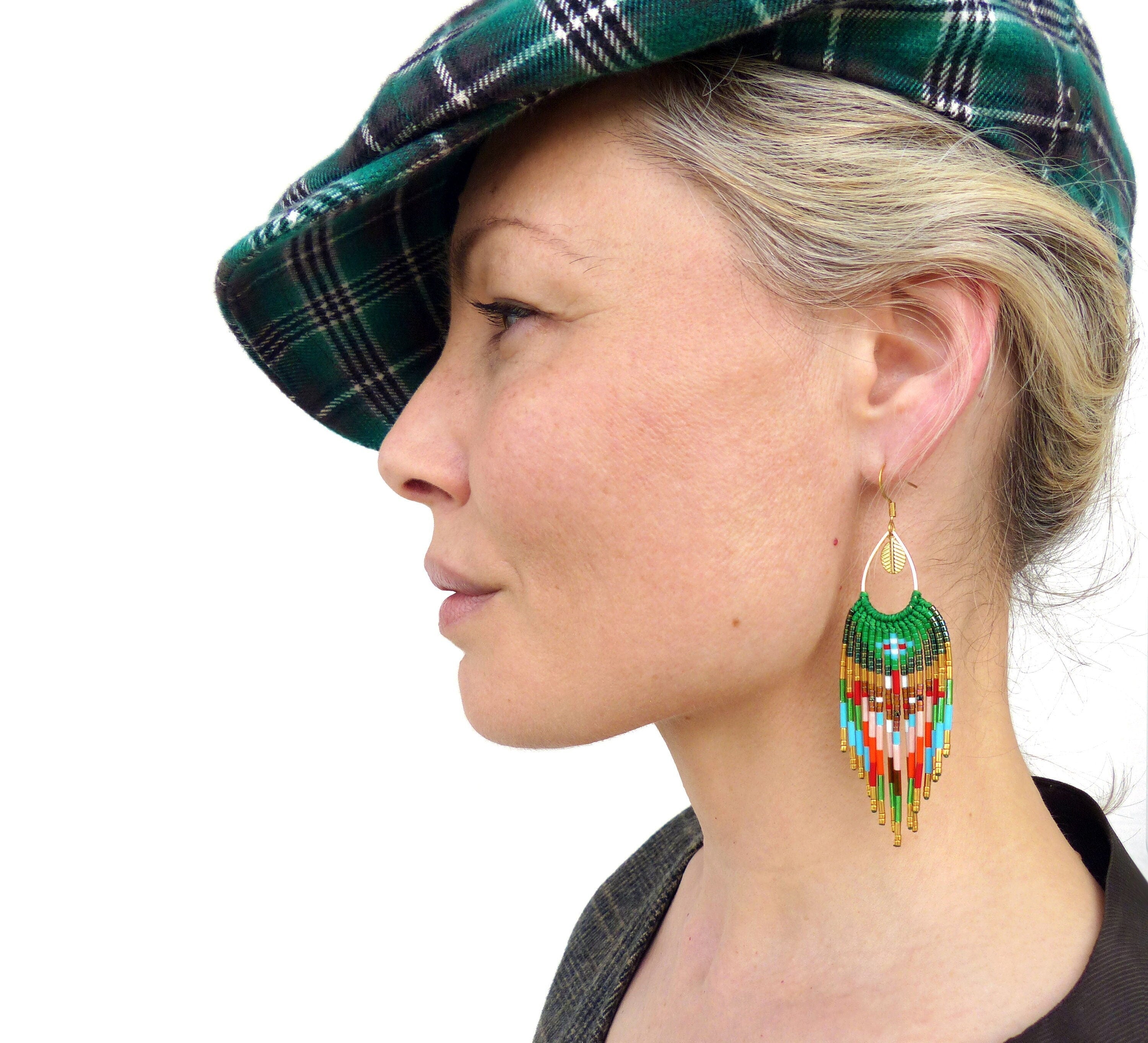 Peut inclure: Une femme portant un chapeau en tartan vert et noir avec de longs cheveux blonds. Elle porte une paire de longues boucles d'oreilles en perles colorées avec des accents dorés.