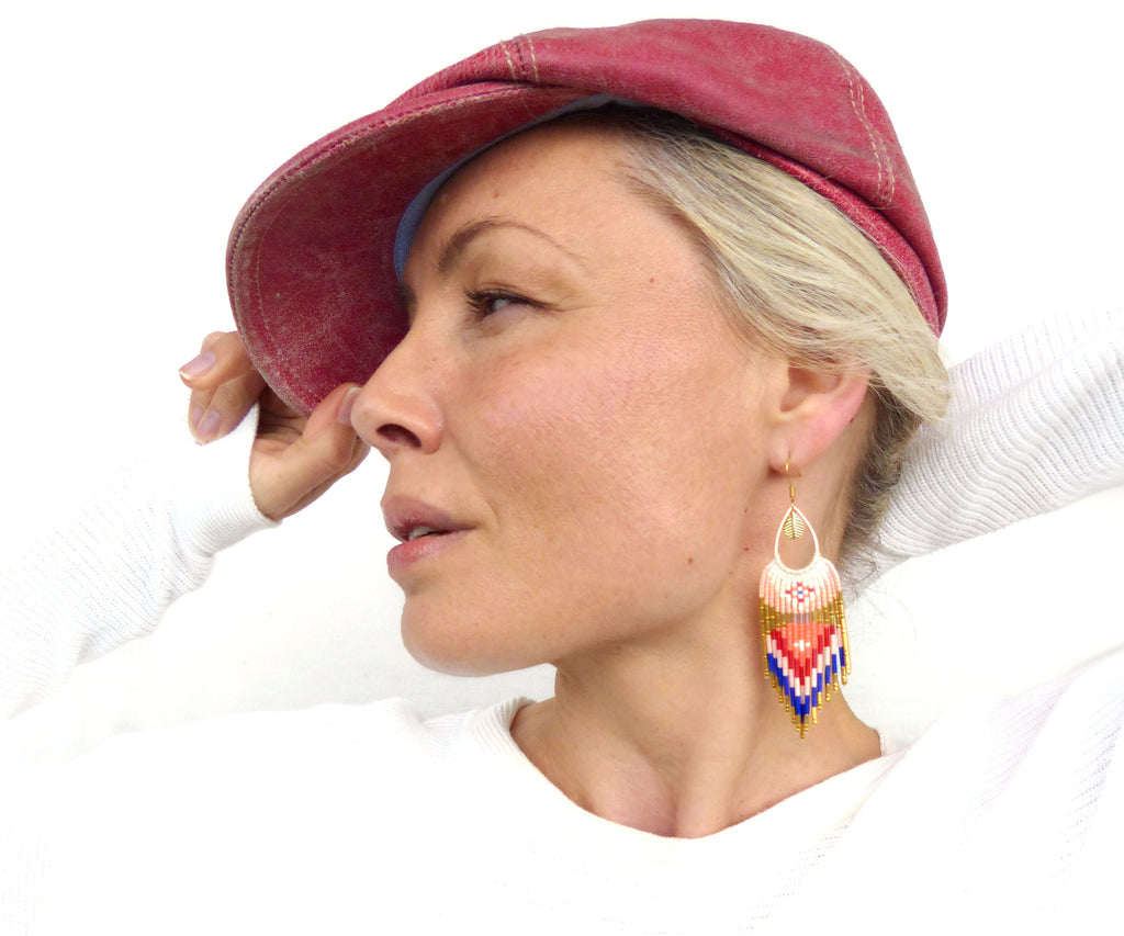 Peut inclure: Une femme portant une casquette de newsboy rouge et un pull blanc à manches longues. Elle porte de longues boucles d'oreilles en perles avec un motif géométrique dans les tons rouge, bleu, blanc et or.