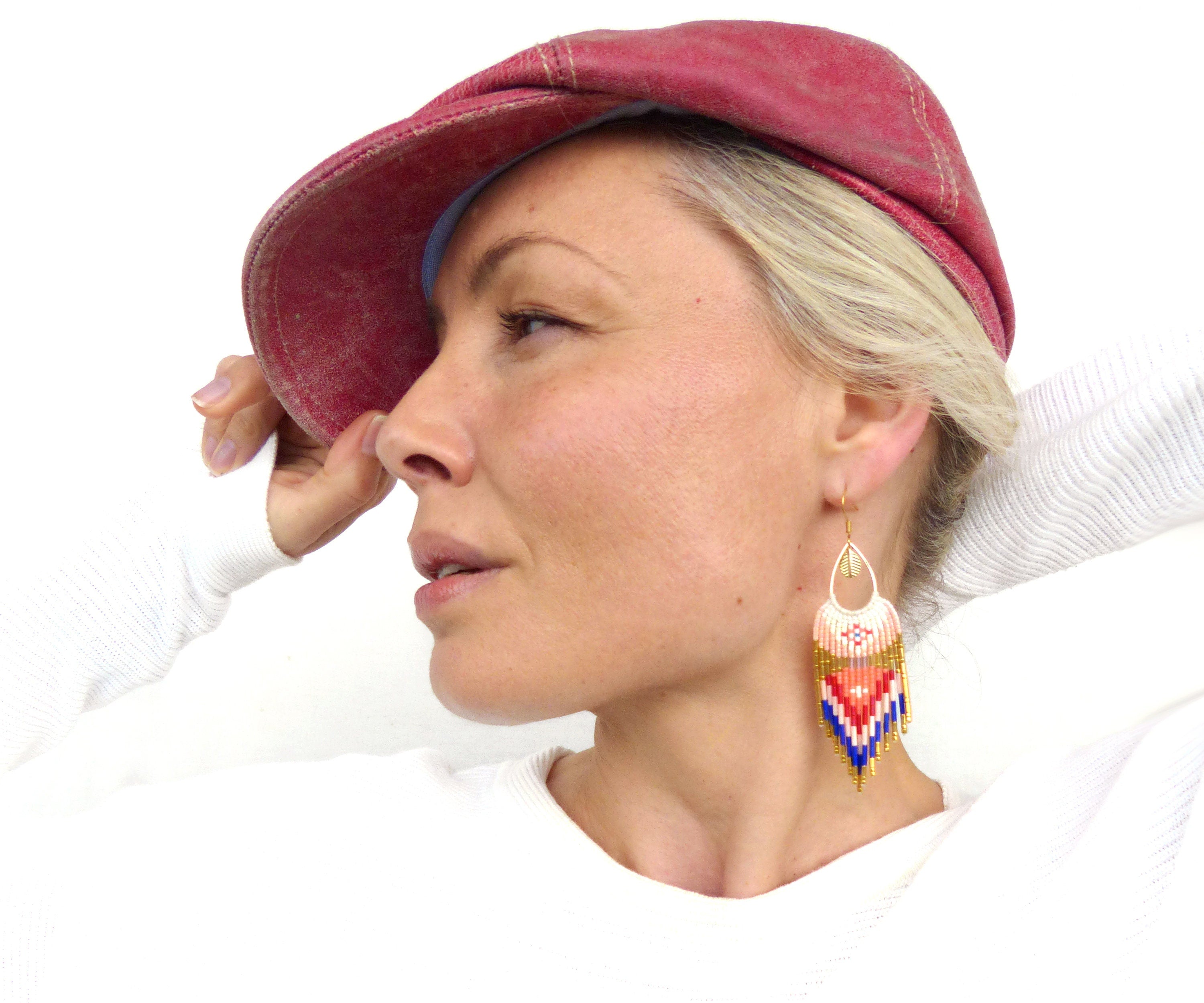 Peut inclure: Une femme portant une casquette de newsboy rouge et un pull blanc à manches longues. Elle porte de longues boucles d'oreilles en perles avec un motif géométrique dans les tons rouge, bleu, blanc et or.