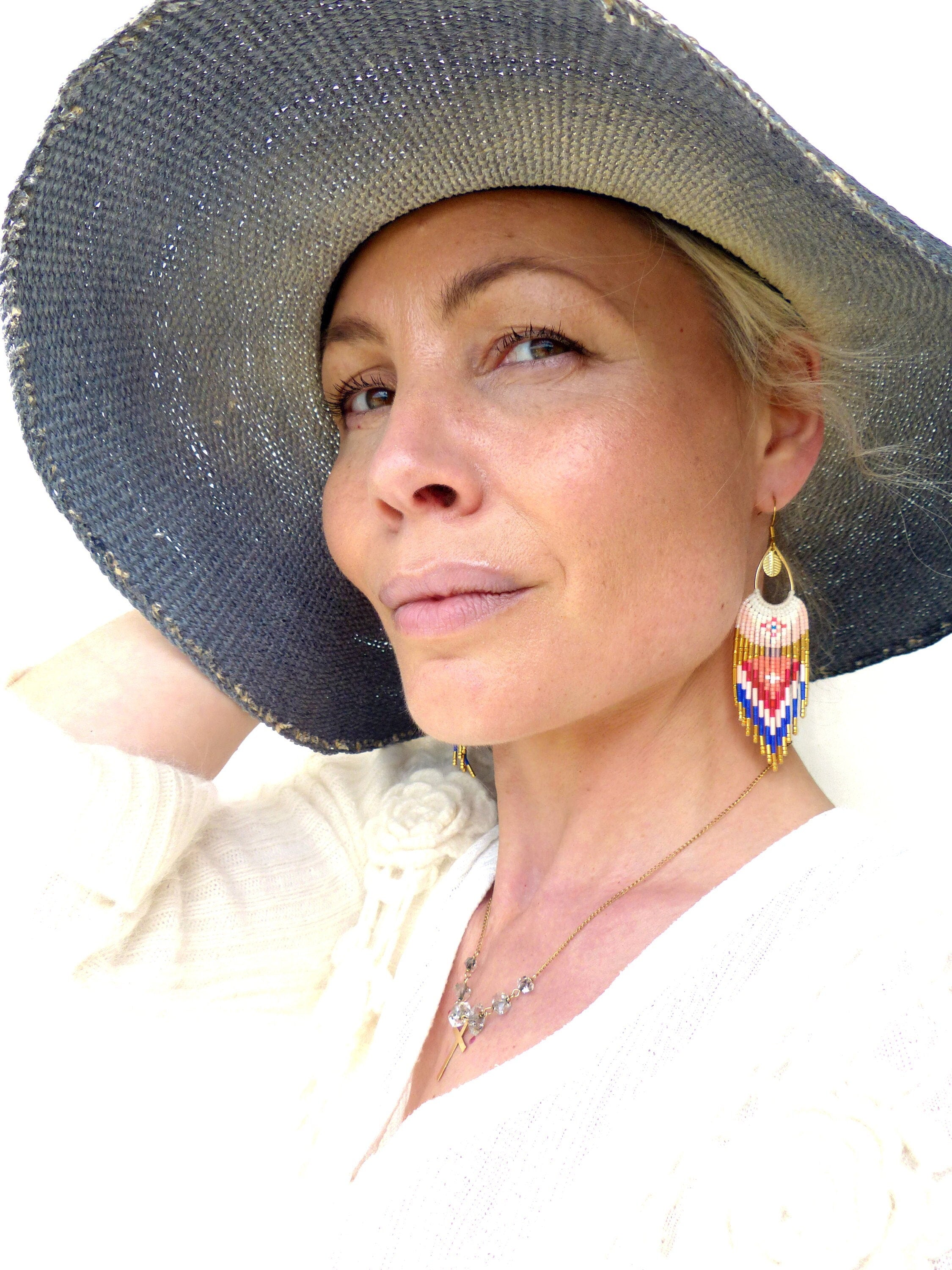 Peut inclure: Une femme portant un chapeau de paille à larges bords avec un haut blanc et un collier en or. Elle porte de longues boucles d'oreilles en perles colorées.