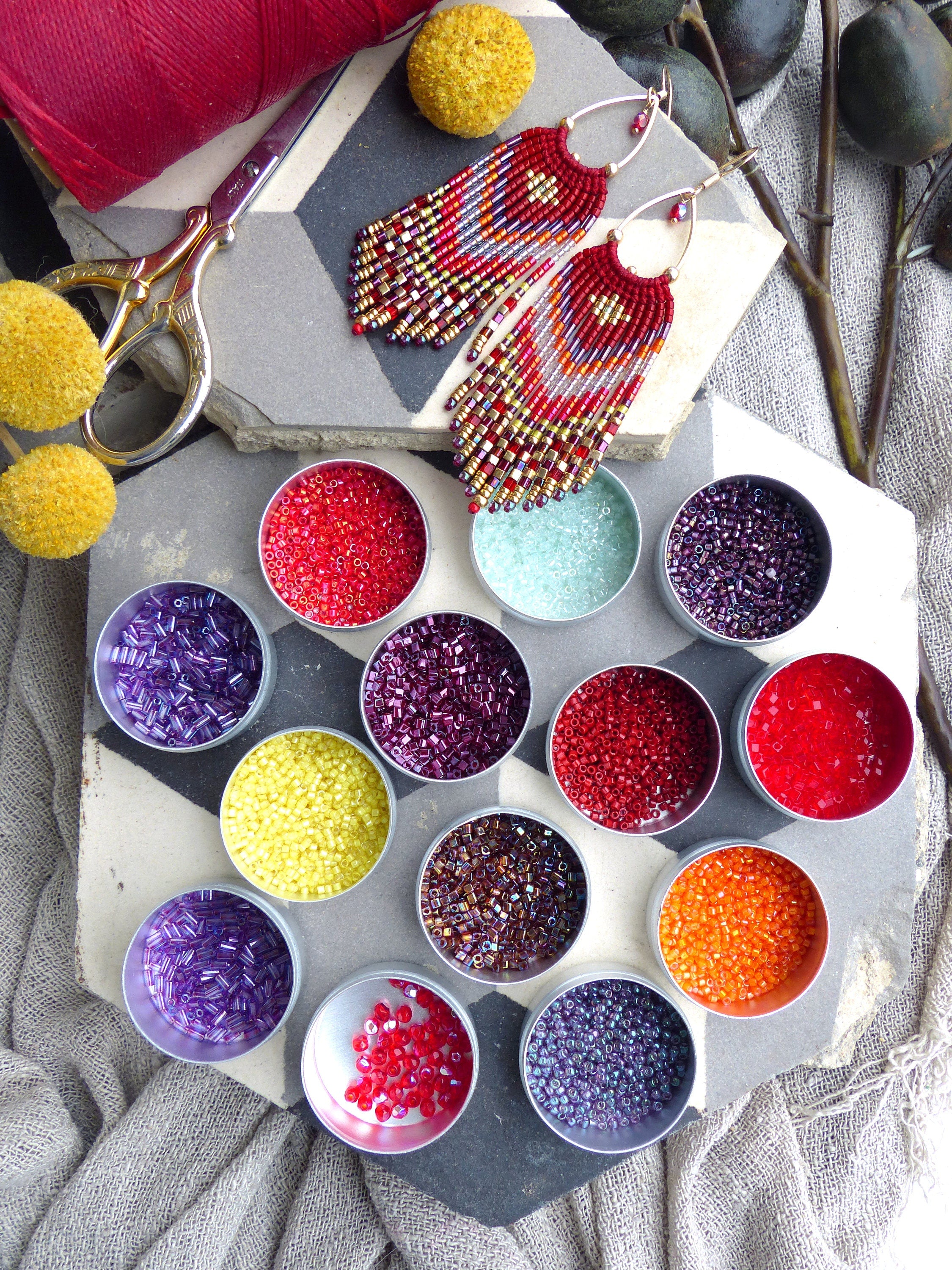 Peut inclure: Assortiment de petits contenants remplis de perles colorées, dont du rouge, de l'orange, du jaune, du violet et du marron. Les perles sont disposées en cercle sur une surface carrelée gris et blanc. Deux paires de boucles d'oreilles en perles avec des perles rouges, orange et violettes pendent au-dessus des contenants.