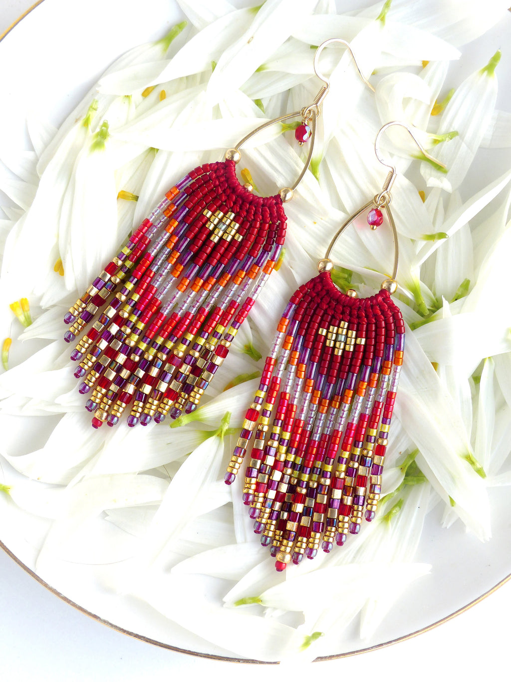 Peut inclure: Une paire de longues boucles d'oreilles en perles avec un motif géométrique rouge, orange, or et violet. Les boucles d'oreilles sont suspendues à des créoles dorées avec des accents de cristal rouge.