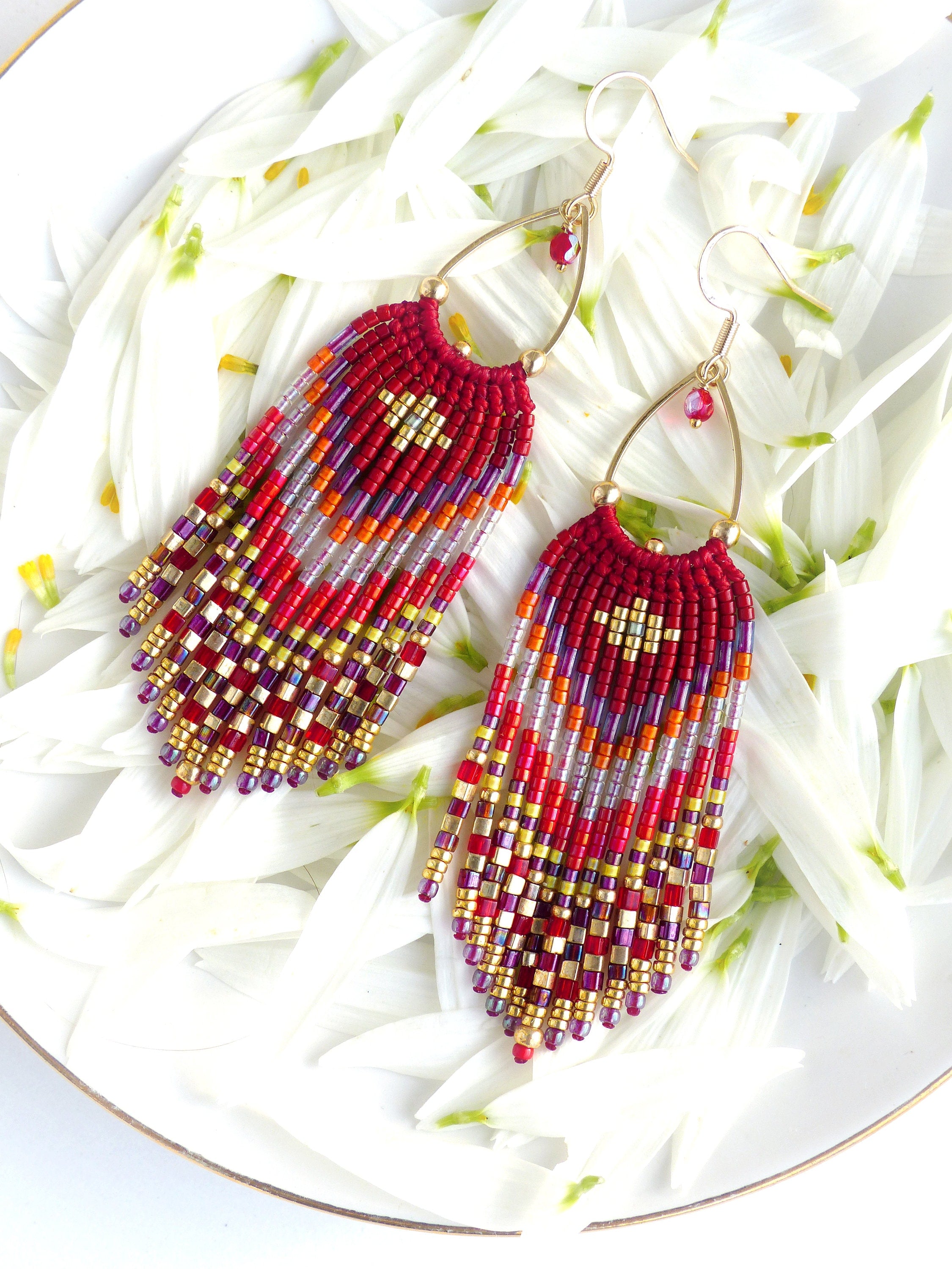 Peut inclure: Une paire de longues boucles d'oreilles en perles avec un motif géométrique rouge, orange, or et violet. Les boucles d'oreilles sont suspendues à des créoles dorées avec des accents de cristal rouge.