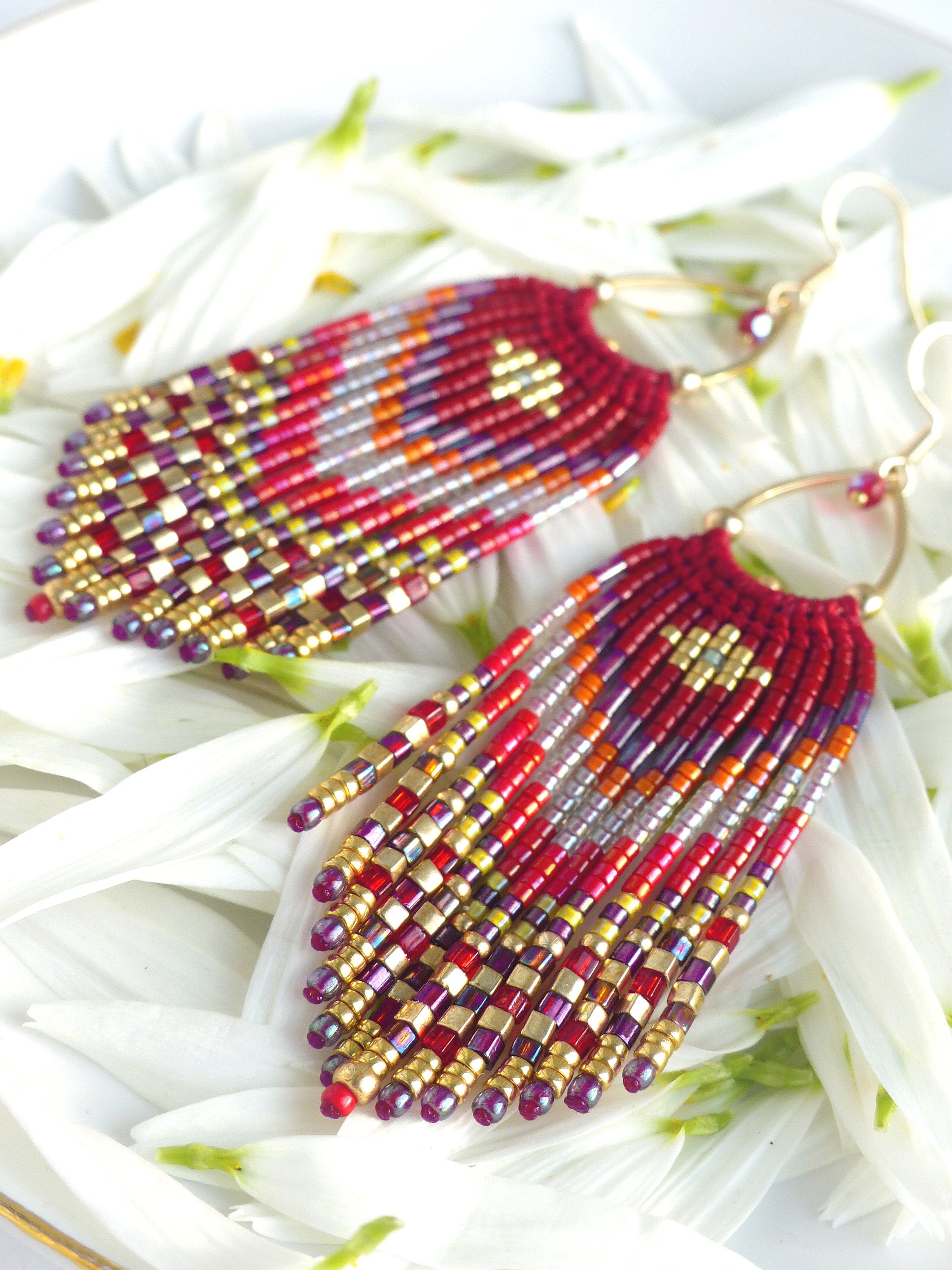 Peut inclure: Une paire de longues boucles d'oreilles en perles avec un motif rouge, or et violet. Les boucles d'oreilles sont faites de petites perles carrées et ont un design en forme de frange.