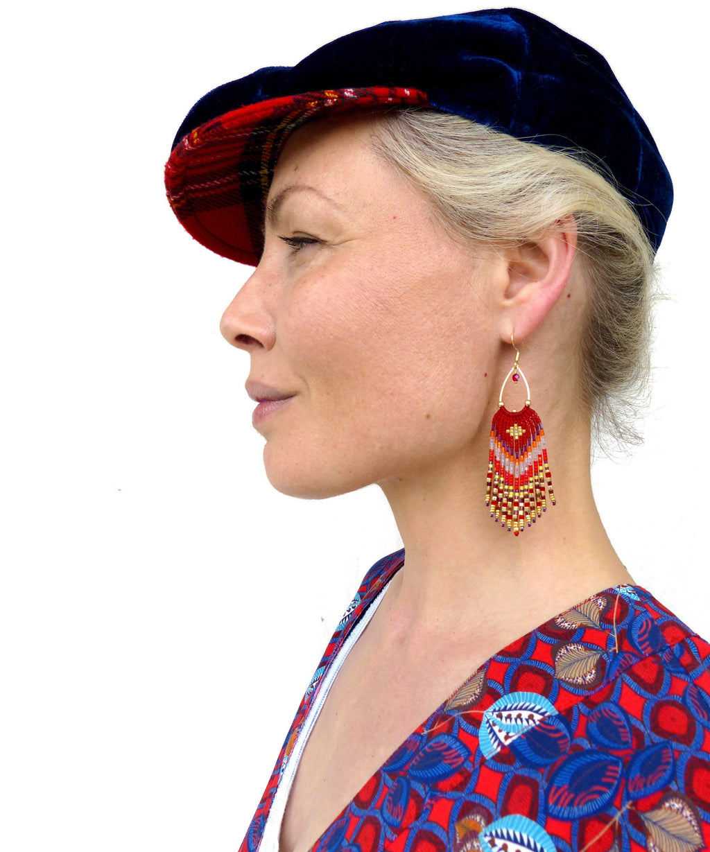 Peut inclure: Une femme portant une casquette de newsboy en tissu écossais bleu et rouge et une chemise à motifs rouge et bleu avec de longues boucles d'oreilles en perles. Les boucles d'oreilles sont rouges, orange et dorées.