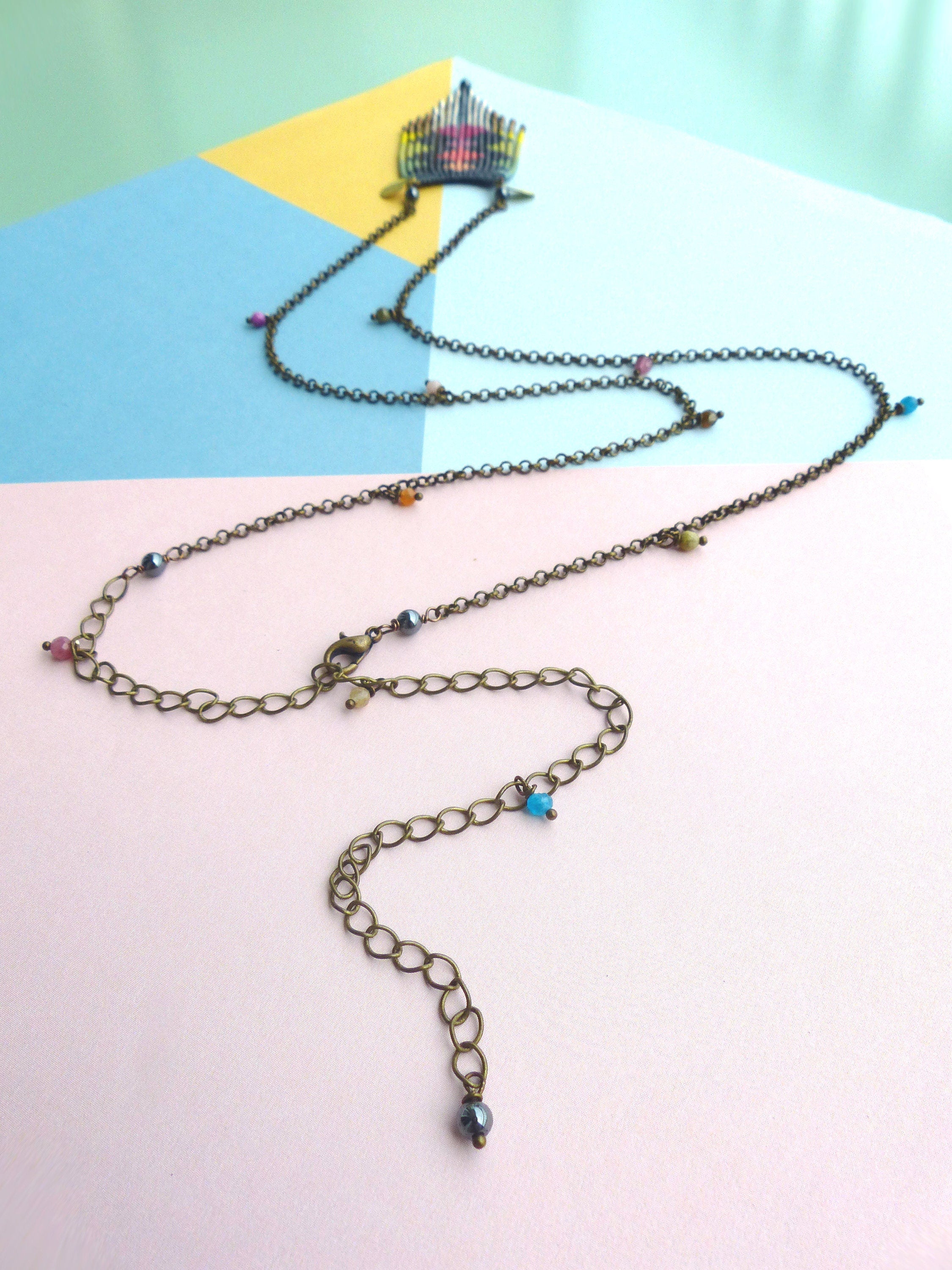 Peut inclure: Un long collier en chaîne bronze avec un pendentif tissé coloré et de petites perles colorées. Le collier a une fermeture à fermoir.