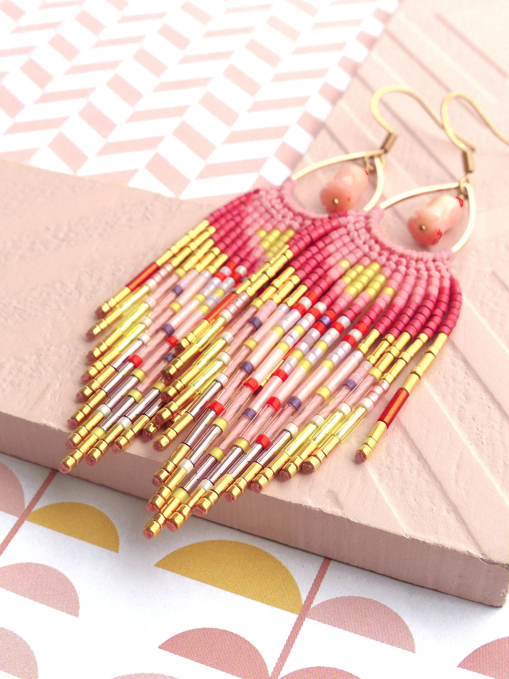 Peut inclure: Une paire de longues boucles d'oreilles en perles avec un schéma de couleurs rose et jaune. Les boucles d'oreilles sont faites de petites perles colorées et d'accents métalliques dorés. Les boucles d'oreilles sont suspendues à des crochets dorés.