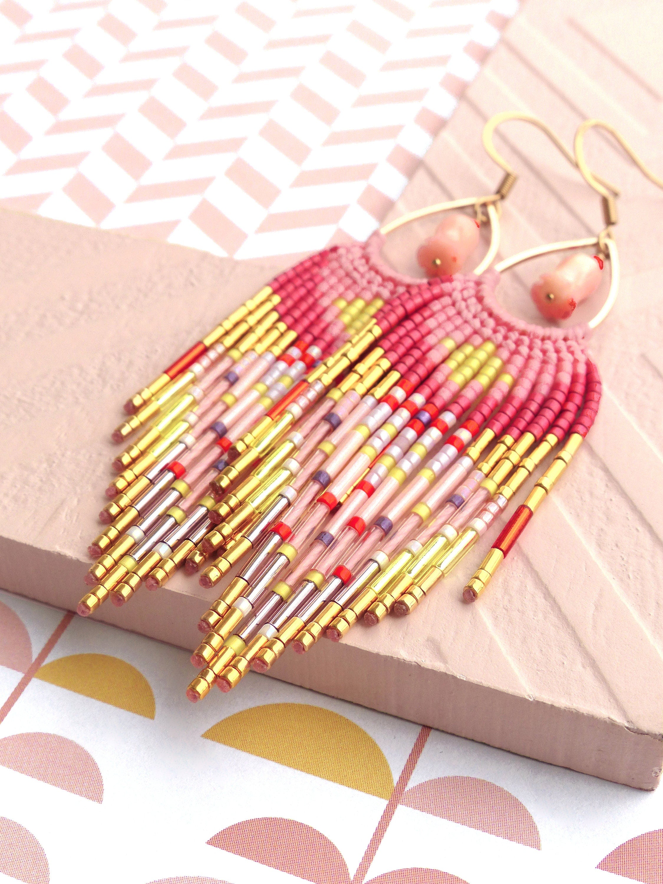 Peut inclure: Une paire de longues boucles d'oreilles en perles avec un schéma de couleurs rose et jaune. Les boucles d'oreilles sont faites de petites perles colorées et d'accents métalliques dorés. Les boucles d'oreilles sont suspendues à des crochets dorés.