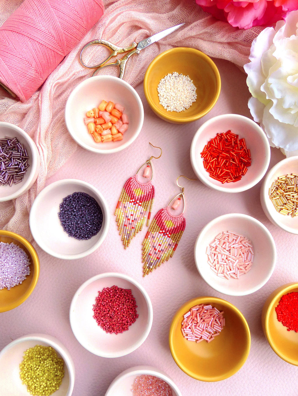 Peut inclure: Une paire de boucles d'oreilles en perles faites à la main avec un design en frange dans des tons de rose, rouge et or. Les boucles d'oreilles sont présentées sur une surface rose avec de petits bols de perles de différentes couleurs.