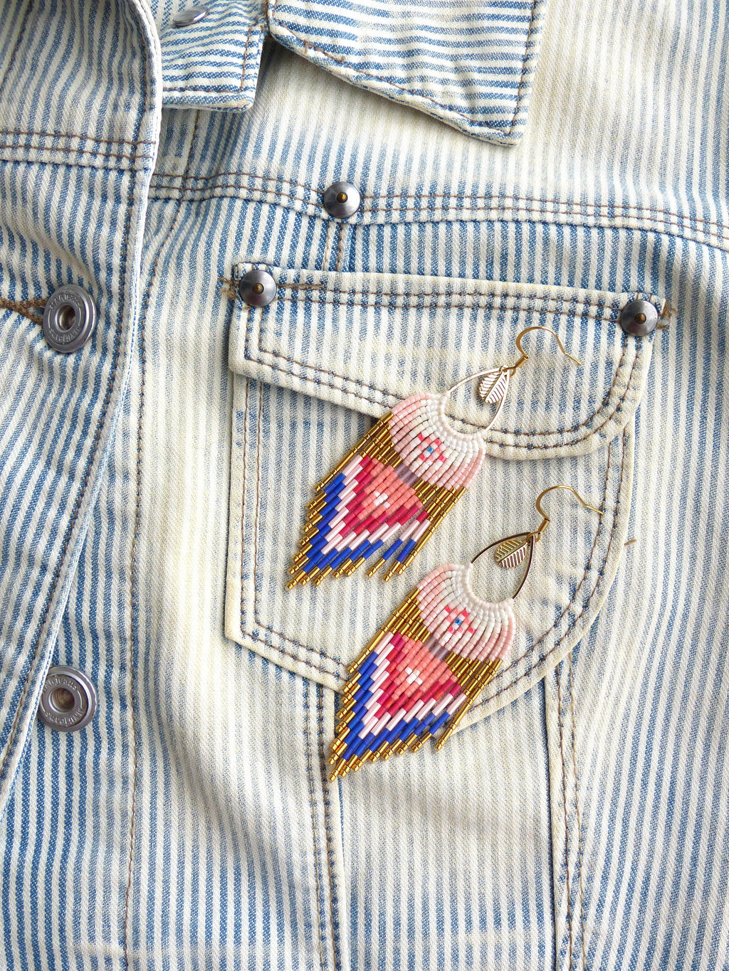 Peut inclure: Une paire de boucles d'oreilles en perles roses, bleues et dorées avec un charme plume suspendu au bas. Les boucles d'oreilles sont suspendues à une veste en jean à rayures blanches.