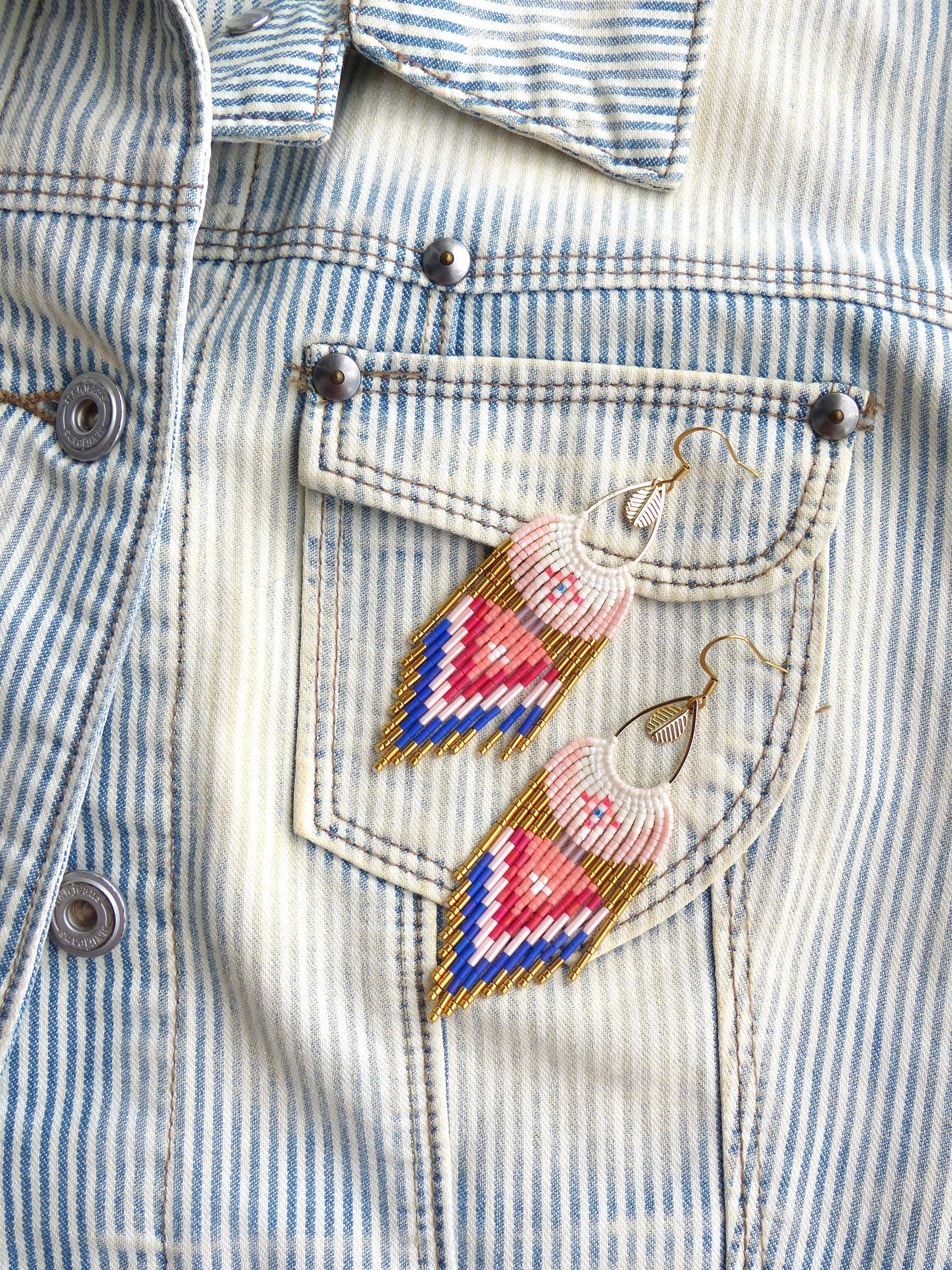 Peut inclure: Une paire de boucles d'oreilles en perles roses, bleues et dorées avec un charme plume suspendu au bas. Les boucles d'oreilles sont suspendues à une veste en jean à rayures blanches.