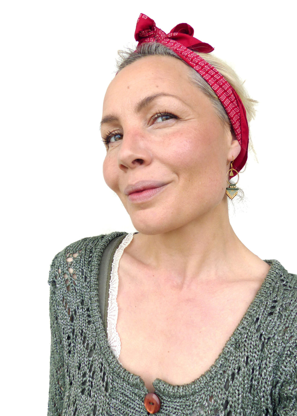 Peut inclure: Une femme aux cheveux courts gris porte un bandana rouge noué autour de sa tête. Elle porte un cardigan en tricot vert avec une fermeture boutonnée. Elle porte également une paire de boucles d'oreilles en or avec des perles vertes et brunes.