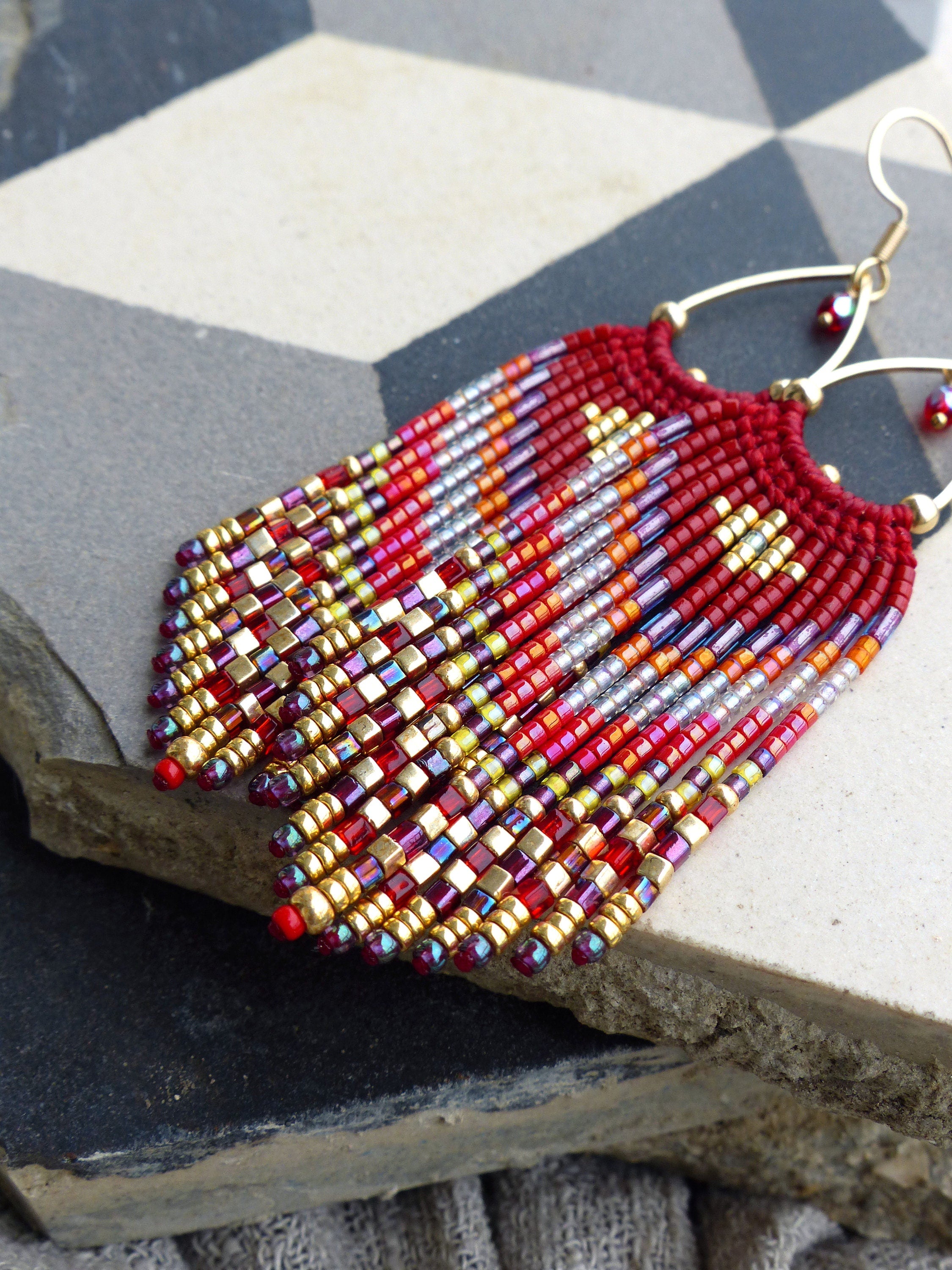 Peut inclure: Boucles d'oreilles en perles rouges, orange, dorées et violettes avec un design en frange. Les boucles d'oreilles sont faites de petites perles carrées et ont un crochet en fil d'or.