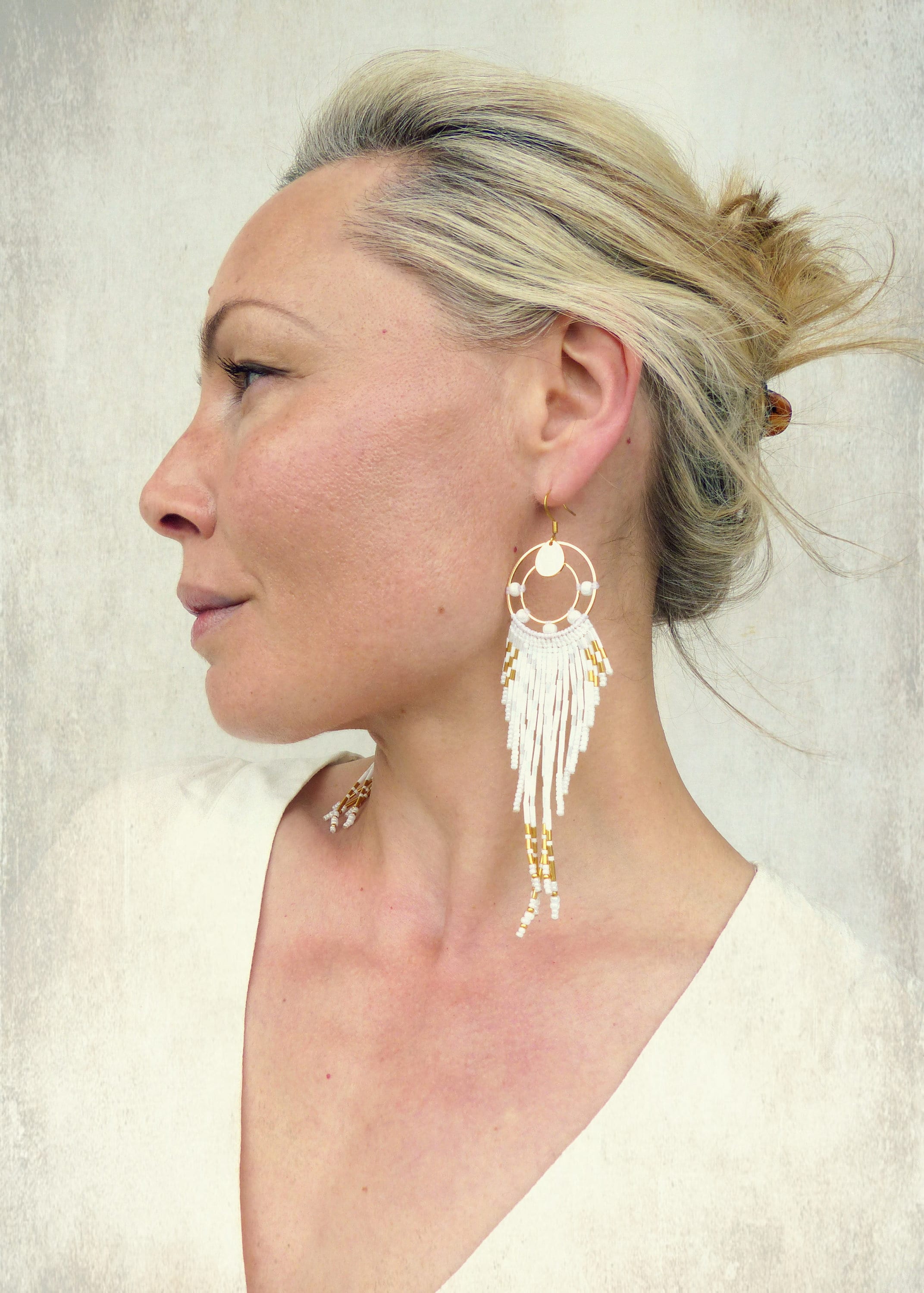 Peut inclure: Boucles d'oreilles créoles dorées avec des franges blanches et dorées. Les boucles d'oreilles présentent un design circulaire avec des perles blanches et de longues franges pendantes blanches et dorées. Un bijou de caractère.