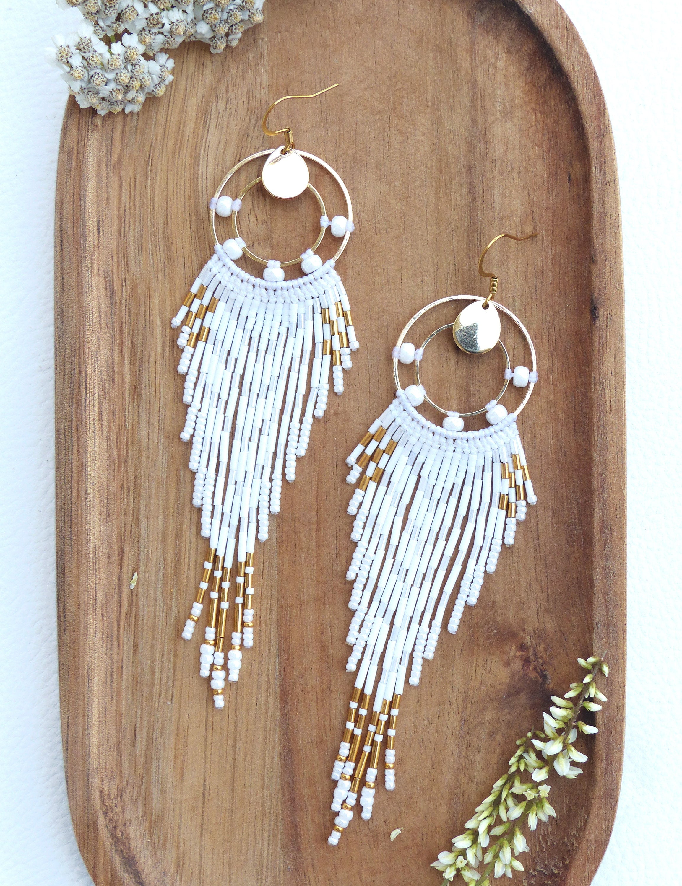 Peut inclure: Une paire de boucles d'oreilles faites à la main avec un design en métal doré. Les boucles d'oreilles présentent un dessus circulaire avec un petit disque doré et une frange de perles blanches avec des accents dorés. Elles sont présentées sur un plateau en bois.