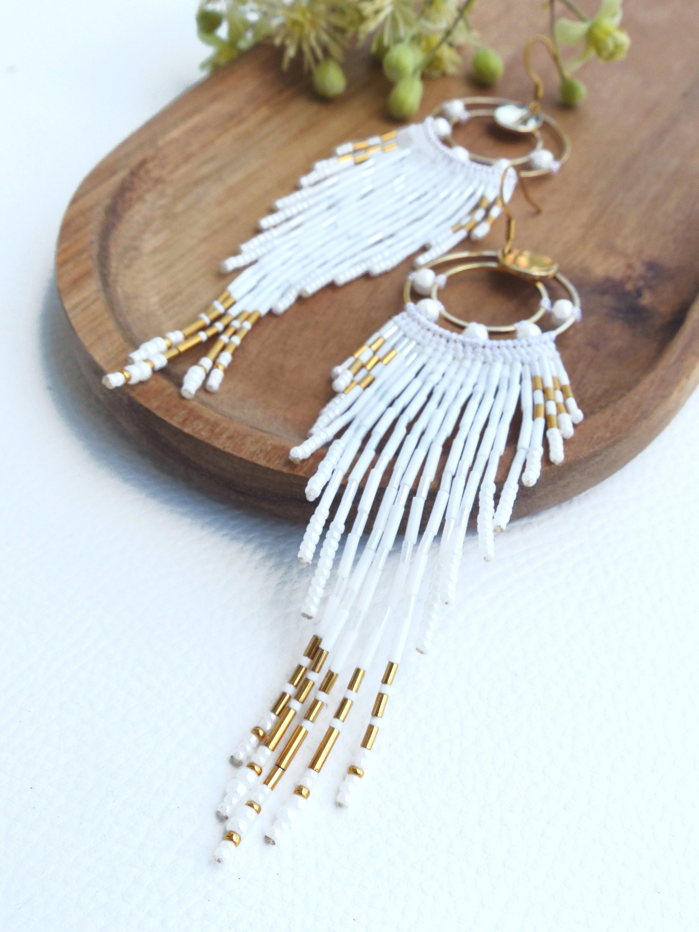 Peut inclure: Une paire de boucles d'oreilles blanches perlées avec des accents dorés. Les boucles d'oreilles présentent un dessus circulaire avec une frange de perles blanches et de perles cylindriques dorées. Les boucles d'oreilles sont présentées sur un plateau en bois.