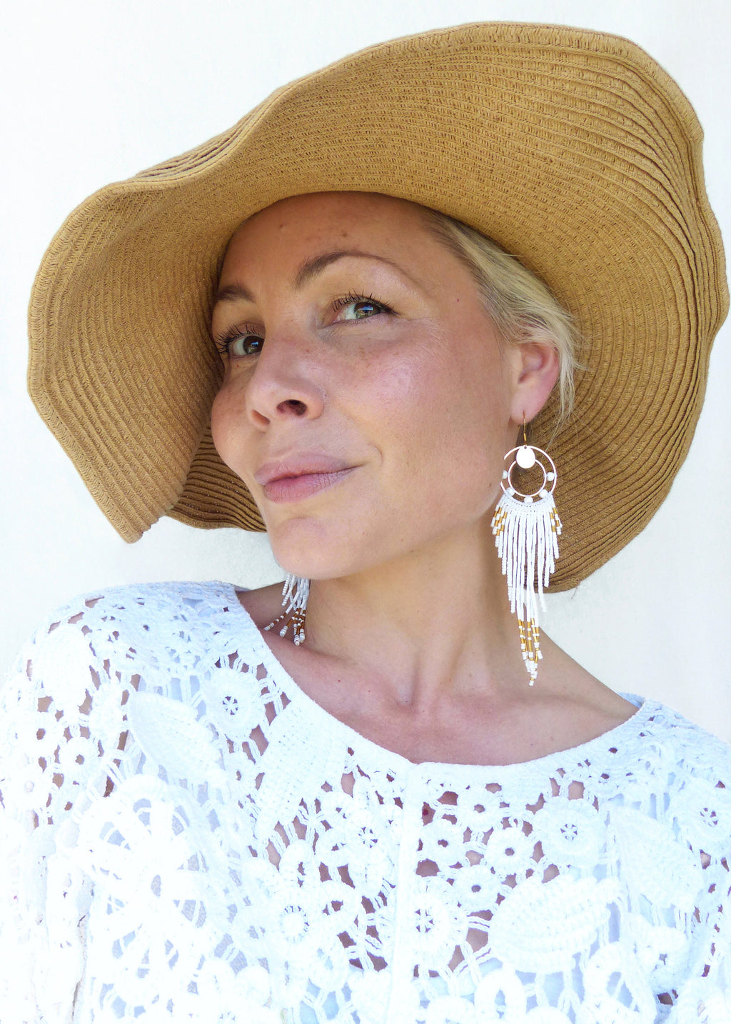 Peut inclure: Une femme porte de grandes boucles d'oreilles blanches à perles avec des accents dorés. Elle porte également un haut en dentelle blanche et un chapeau de paille à larges bords. Les boucles d'oreilles ont un design circulaire.