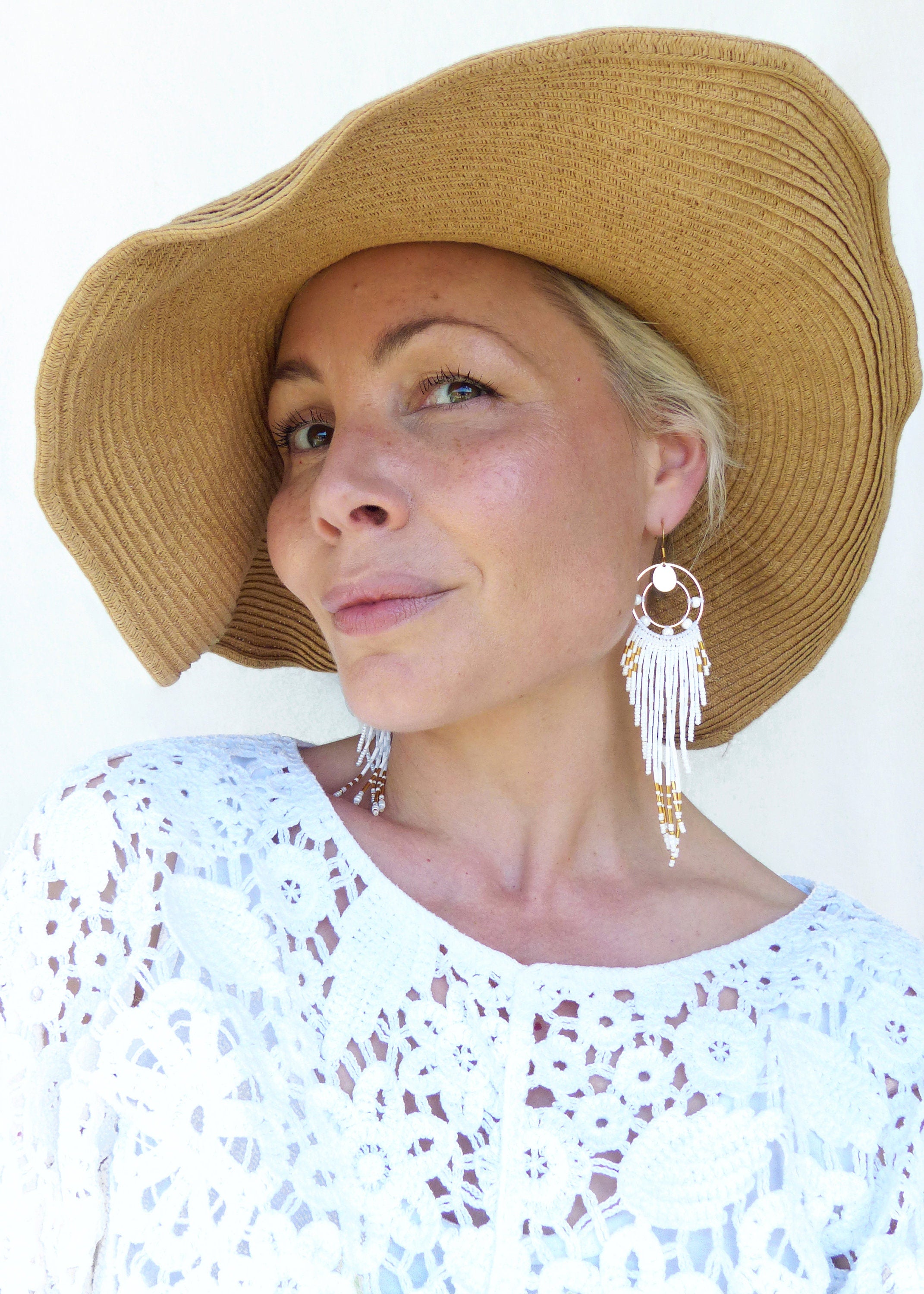 Peut inclure: Une femme porte de grandes boucles d'oreilles blanches à perles avec des accents dorés. Elle porte également un haut en dentelle blanche et un chapeau de paille à larges bords. Les boucles d'oreilles ont un design circulaire.