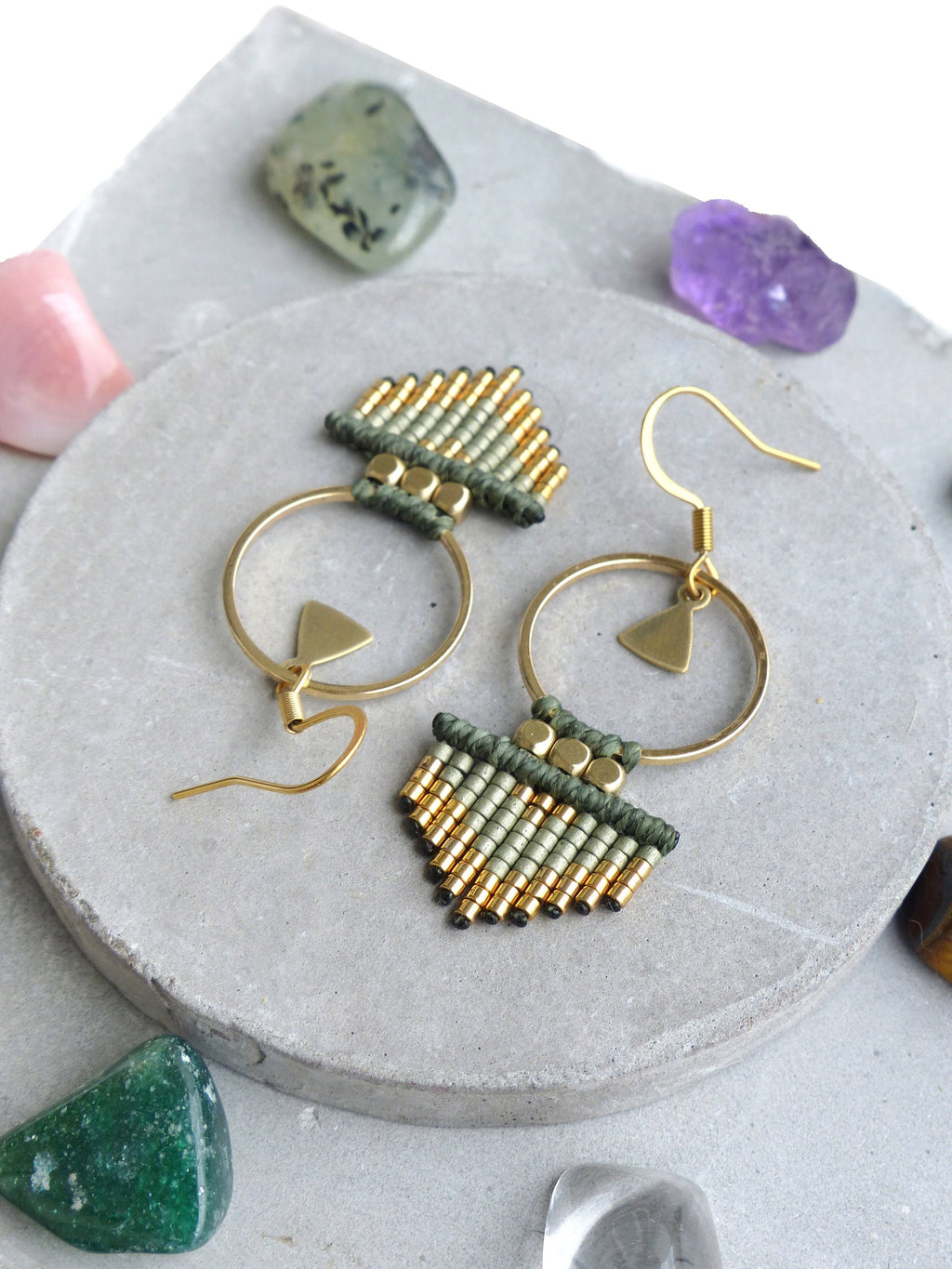 Peut inclure: Une paire de boucles d'oreilles créoles dorées avec des franges de perles vertes et dorées. Les boucles d'oreilles sont sur une surface grise avec différentes pierres.