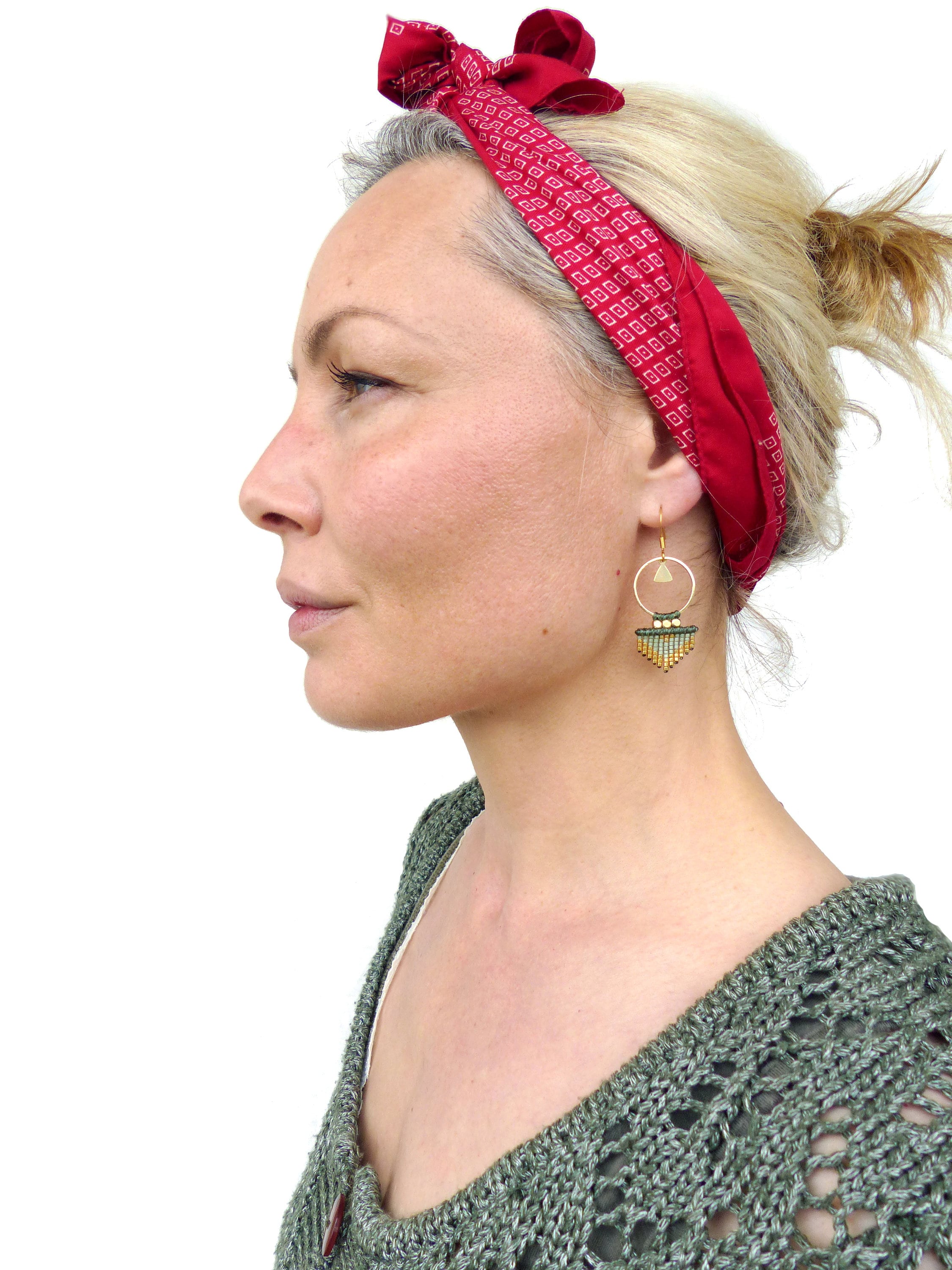 Peut inclure: Une femme portant un bandana rouge à motif géométrique blanc et un pull en tricot vert. Elle porte des boucles d'oreilles créoles dorées avec un pendentif en perles vertes et dorées.