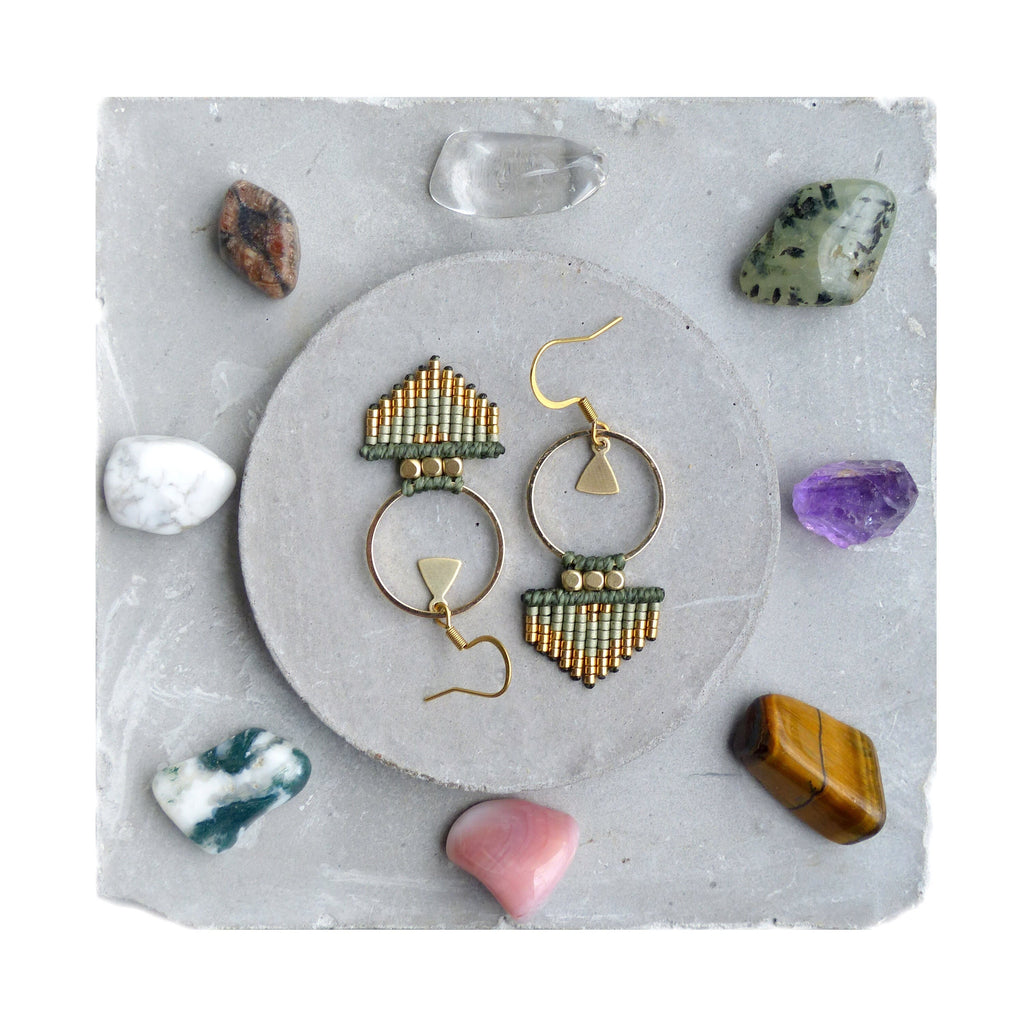 Peut inclure: Une paire de boucles d'oreilles en perles dorées et vertes avec un motif géométrique. Les boucles d'oreilles sont suspendues à une surface grise avec diverses pierres.