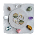 Peut inclure: Une paire de boucles d'oreilles en perles dorées et vertes avec un motif géométrique. Les boucles d'oreilles sont suspendues à une surface grise avec diverses pierres.