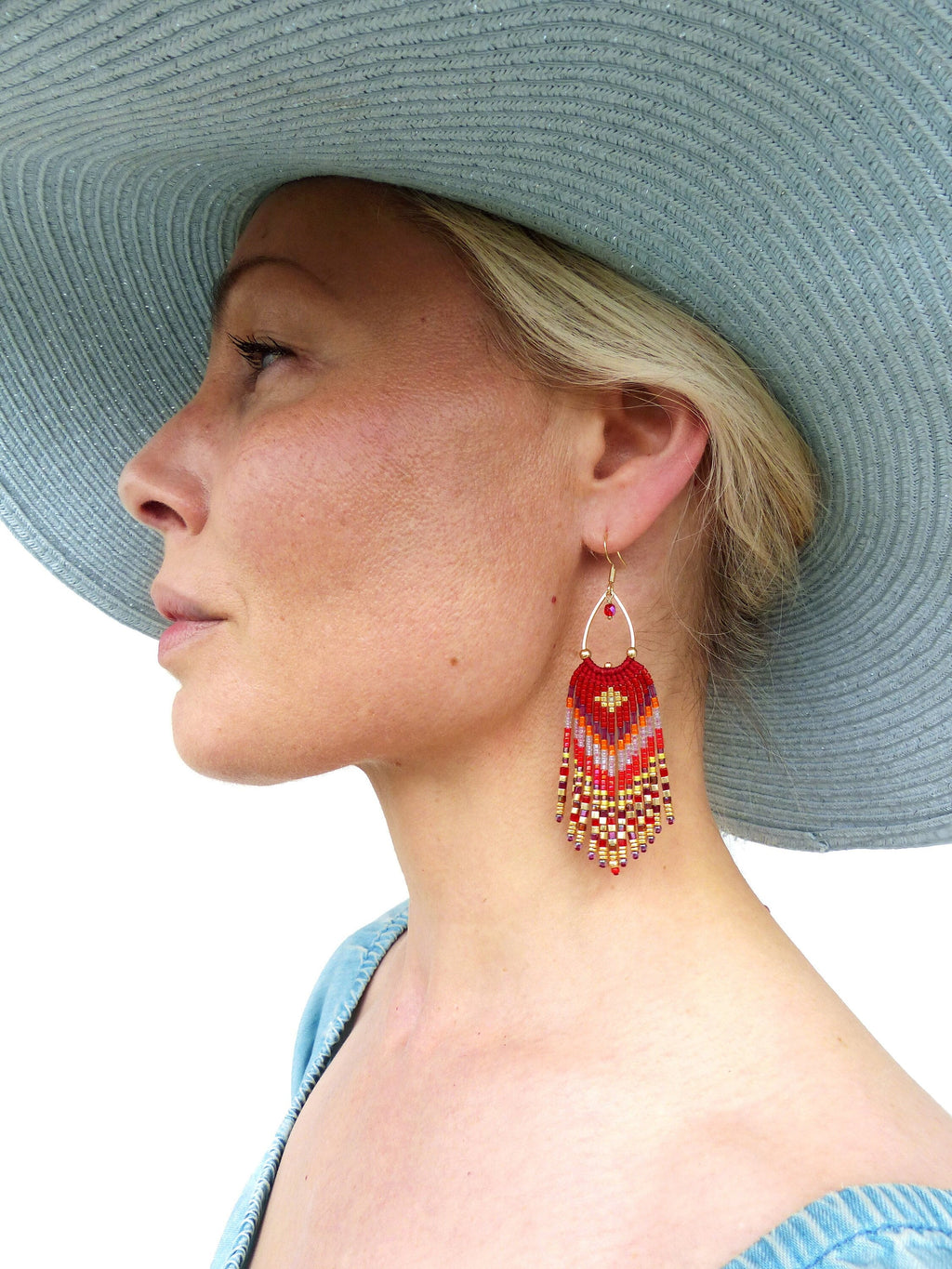 Peut inclure: Une paire de boucles d'oreilles en perles rouges, dorées et blanches avec un design en frange. Les boucles d'oreilles pendent de l'oreille d'une personne portant un chapeau bleu.