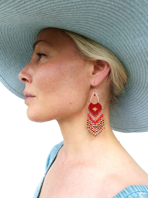 Peut inclure: Une paire de boucles d'oreilles en perles rouges, dorées et blanches avec un design en frange. Les boucles d'oreilles pendent de l'oreille d'une personne portant un chapeau bleu.