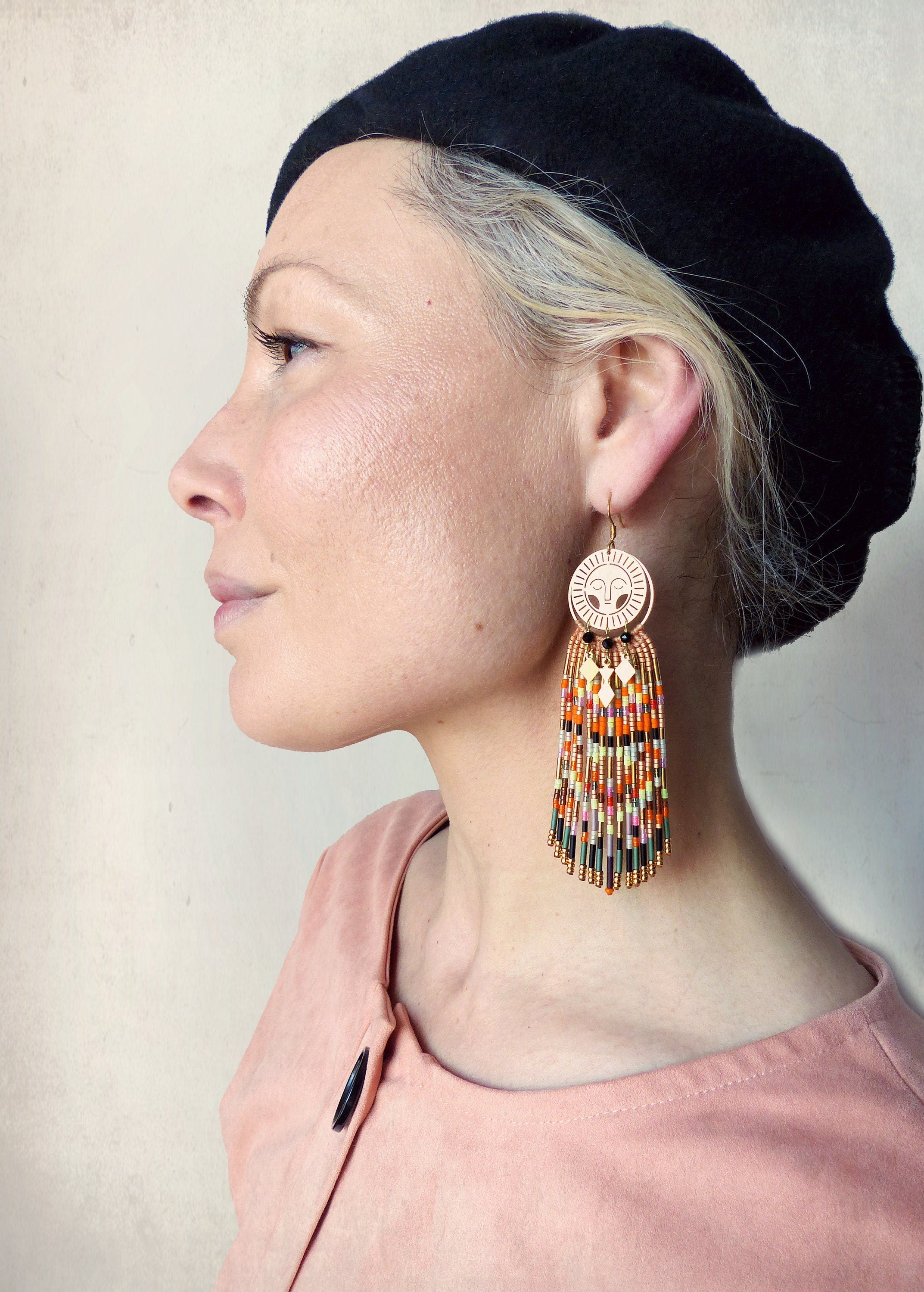 Peut inclure: Une paire de longues boucles d'oreilles en perles colorées avec un charme en forme de soleil doré. Les boucles d'oreilles pendent de l'oreille d'une personne portant un béret noir.
