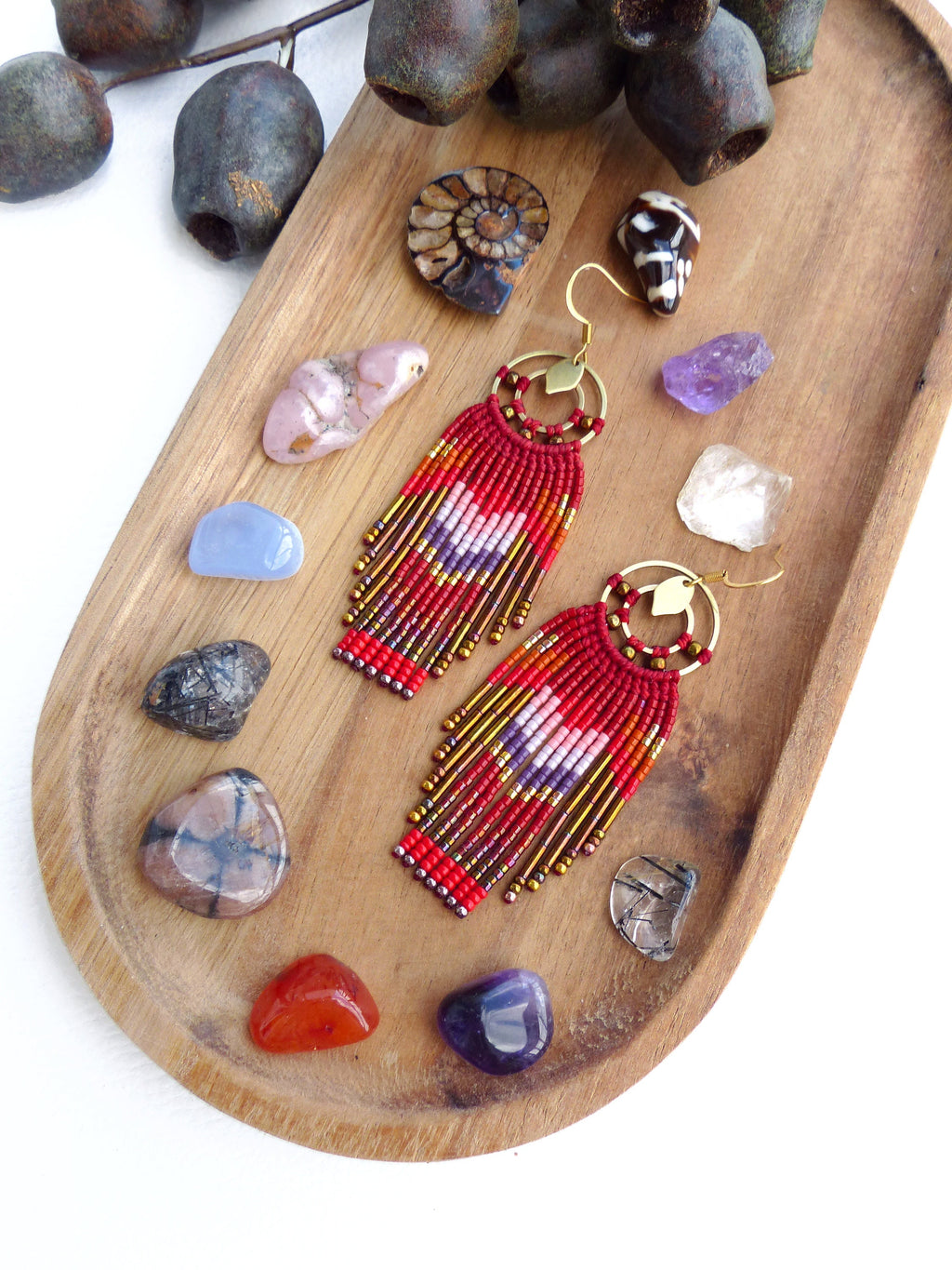 Peut inclure: Une paire de boucles d'oreilles en perles rouges, dorées et blanches avec un design en frange. Les boucles d'oreilles sont suspendues sur une surface en bois avec diverses pierres et cristaux.