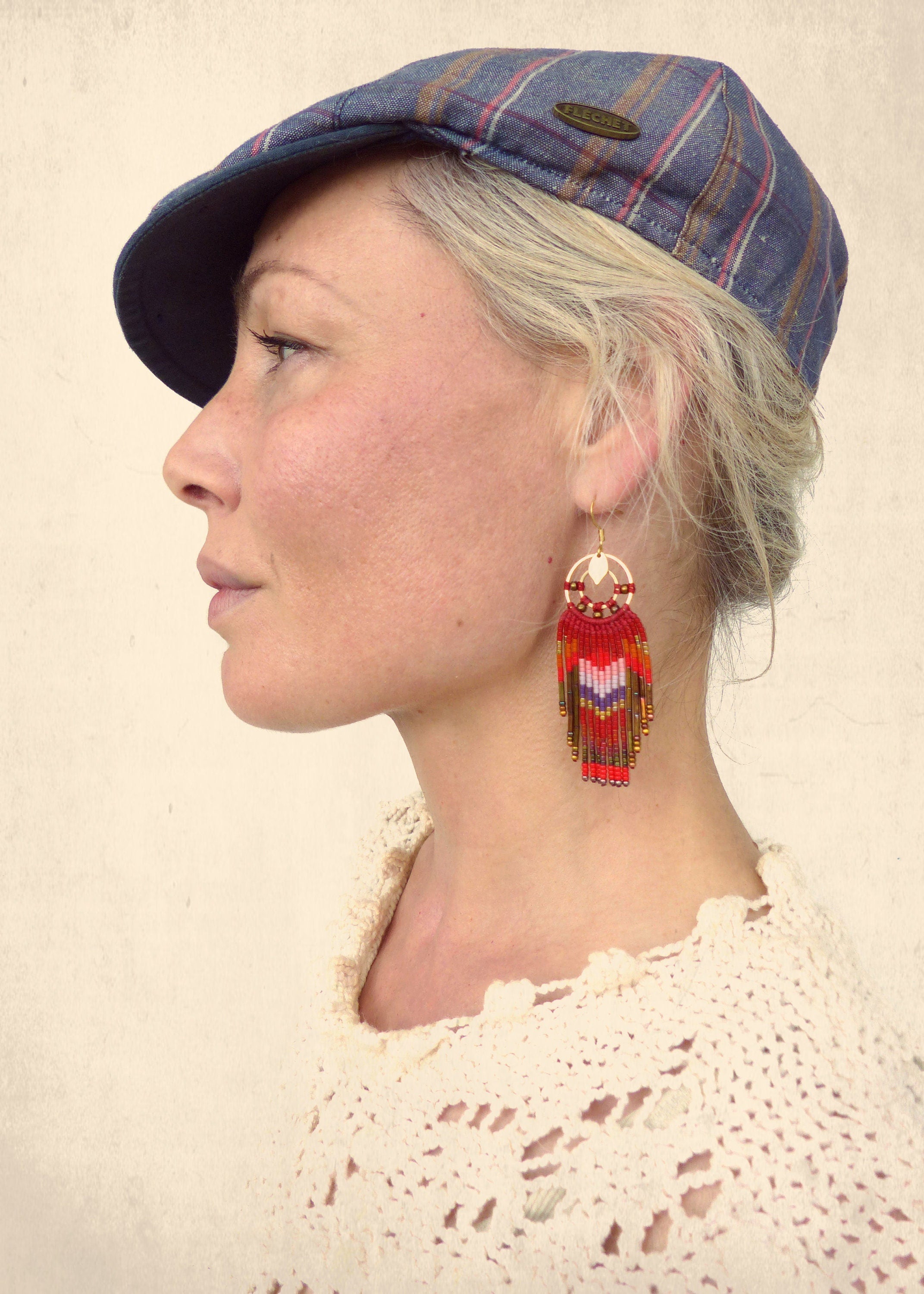 Peut inclure: Une femme portant une casquette de newsboy bleu et blanc à carreaux avec des boucles d'oreilles en perles rouges et blanches. Les boucles d'oreilles sont longues et pendantes, avec un motif géométrique.