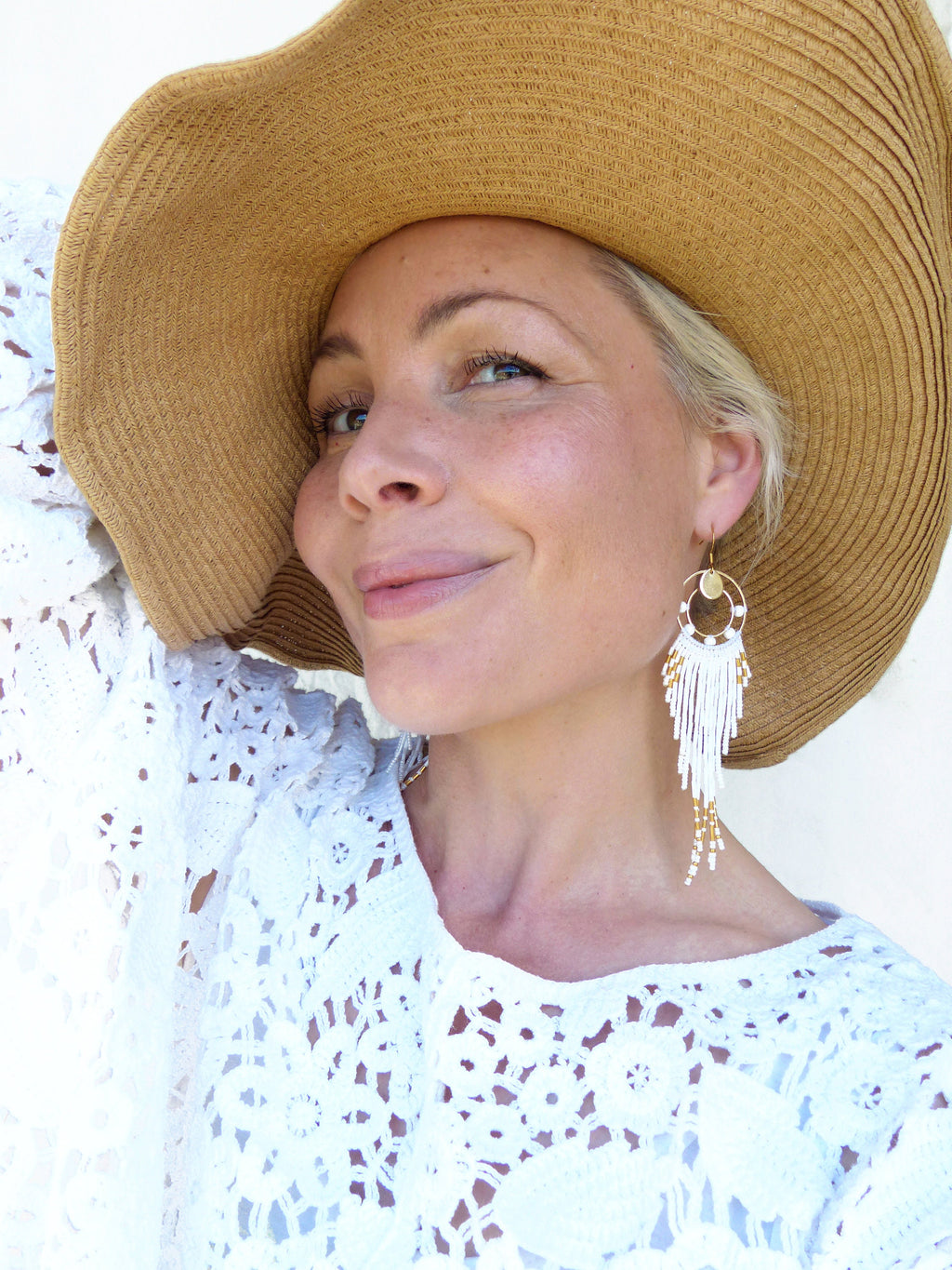 Peut inclure: Une femme porte un chapeau de paille à larges bords et un haut en dentelle blanche. L'accent est mis sur une paire de longues boucles d'oreilles pendantes avec des perles blanches et dorées. Les boucles d'oreilles ont un design circulaire en haut.