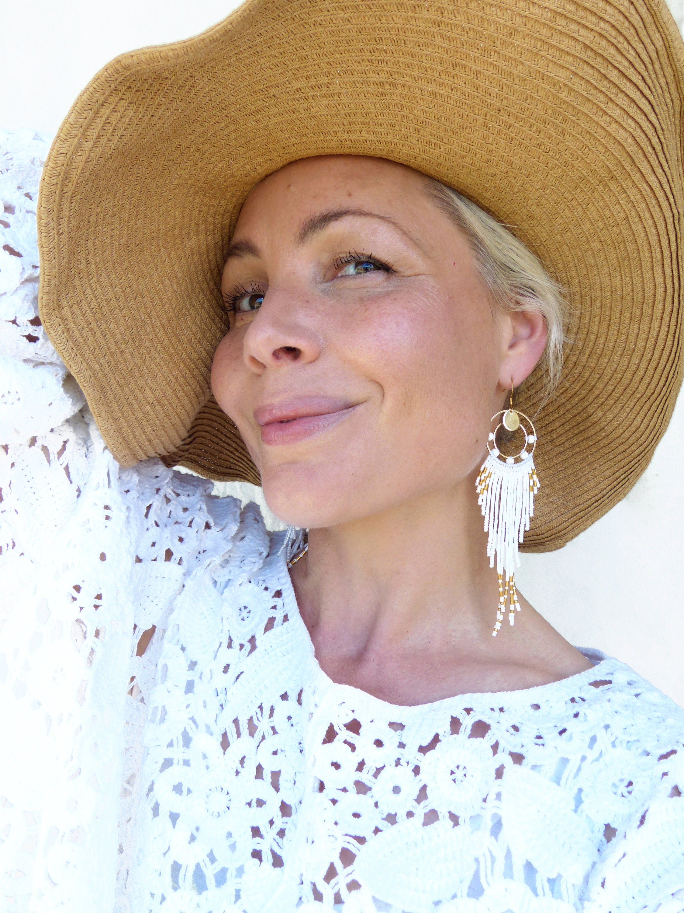 Peut inclure: Une femme porte un chapeau de paille à larges bords et un haut en dentelle blanche. L'accent est mis sur une paire de longues boucles d'oreilles pendantes avec des perles blanches et dorées. Les boucles d'oreilles ont un design circulaire en haut.