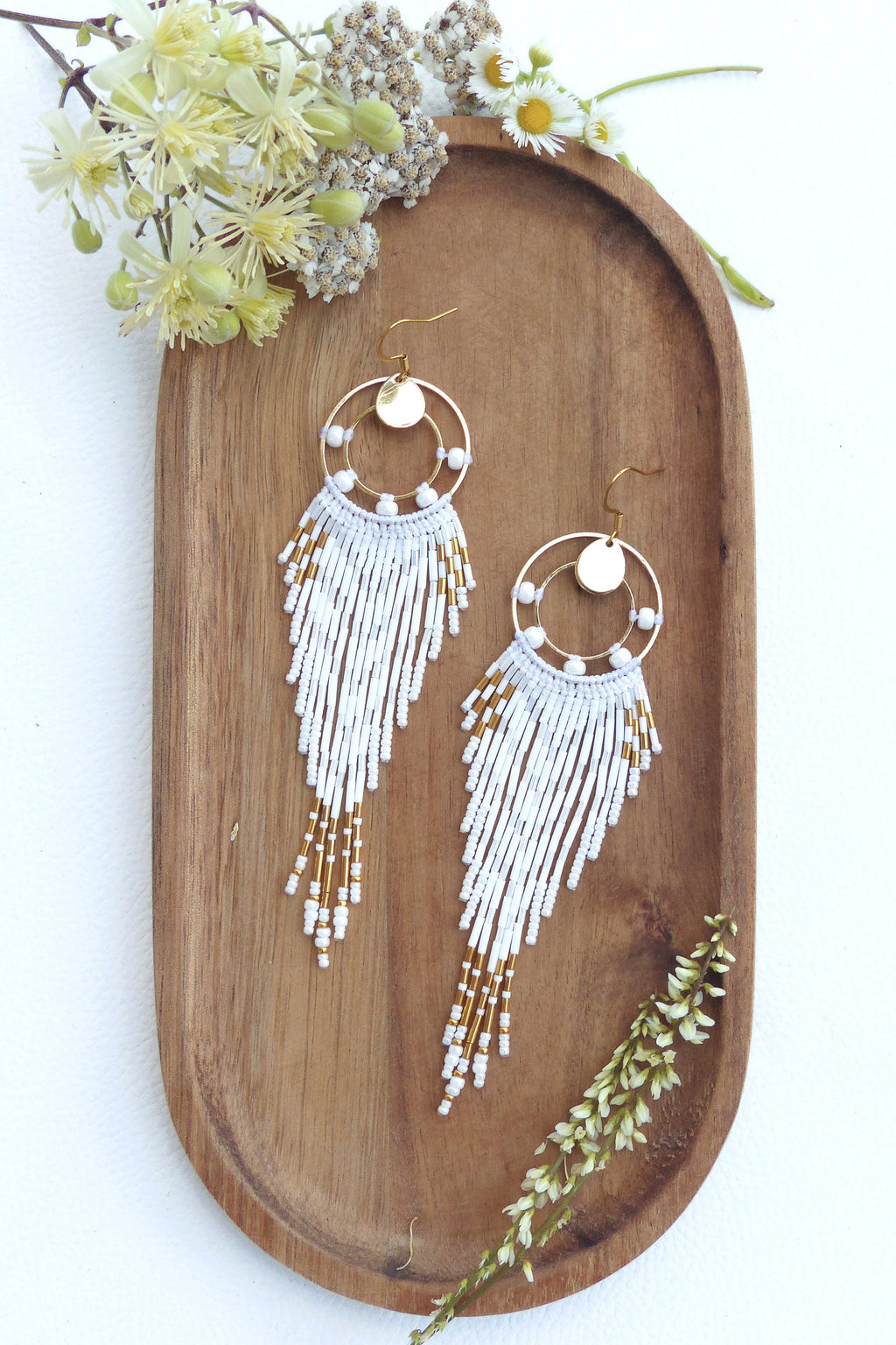 Peut inclure: Paire de boucles d'oreilles faites à la main avec un design circulaire doré, des perles blanches et des accents dorés. Les boucles d'oreilles sont présentées sur un plateau en bois avec des éléments floraux. Elles ont un design à franges.