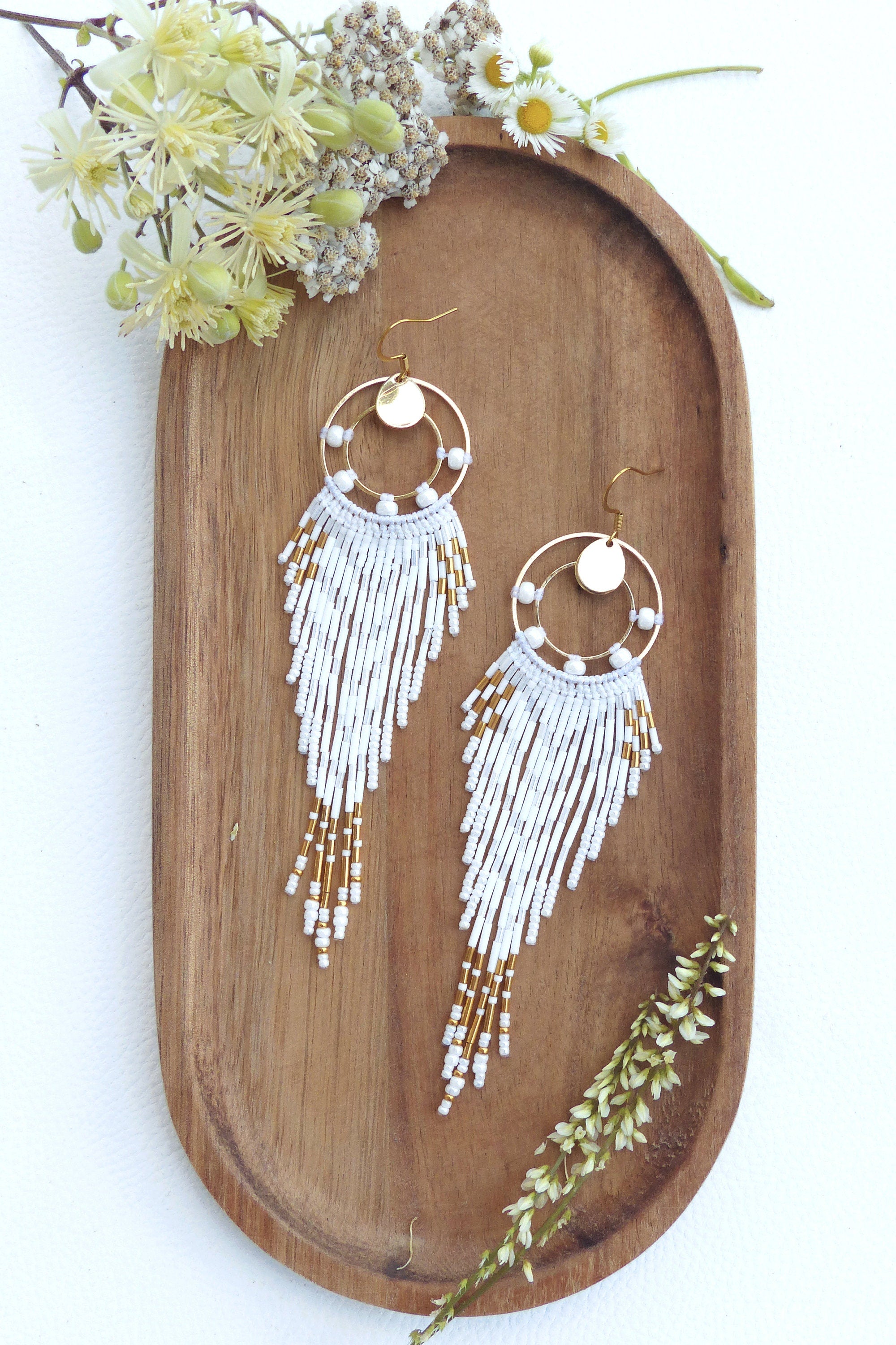 Peut inclure: Paire de boucles d'oreilles faites à la main avec un design circulaire doré, des perles blanches et des accents dorés. Les boucles d'oreilles sont présentées sur un plateau en bois avec des éléments floraux. Elles ont un design à franges.