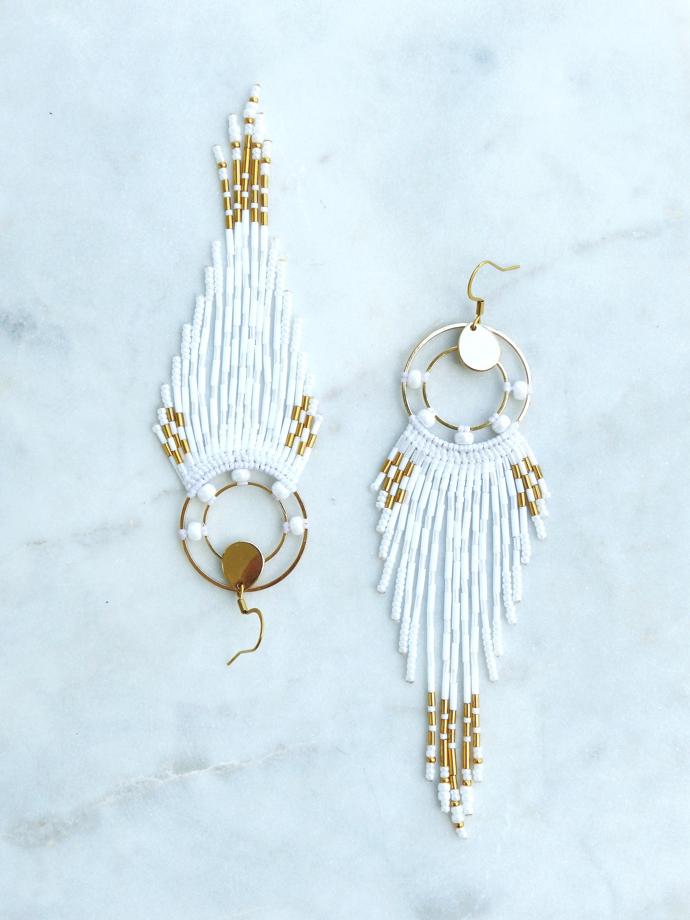 Peut inclure: Une paire de boucles d'oreilles en perles blanches avec des accents dorés. Les boucles d'oreilles présentent un design à franges avec un cadre doré circulaire et un petit disque doré. Fermeture à crochet.