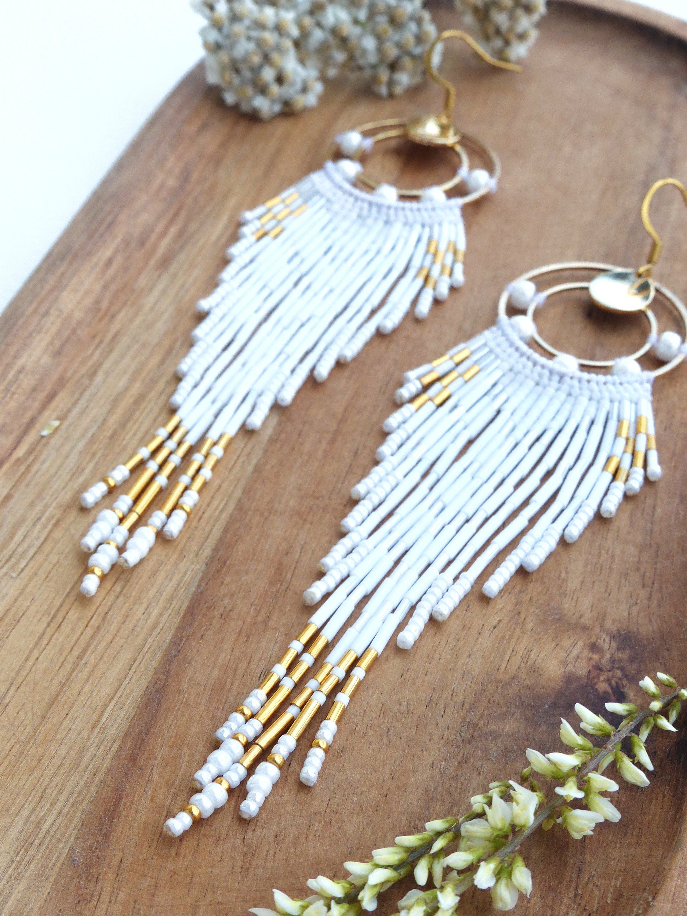 Peut inclure: Une paire de boucles d'oreilles à franges blanches perlées avec des accents dorés. Les boucles d'oreilles ont un dessus doré circulaire avec des perles blanches et une longue frange de perles blanches avec des détails de perles dorées et blanches.